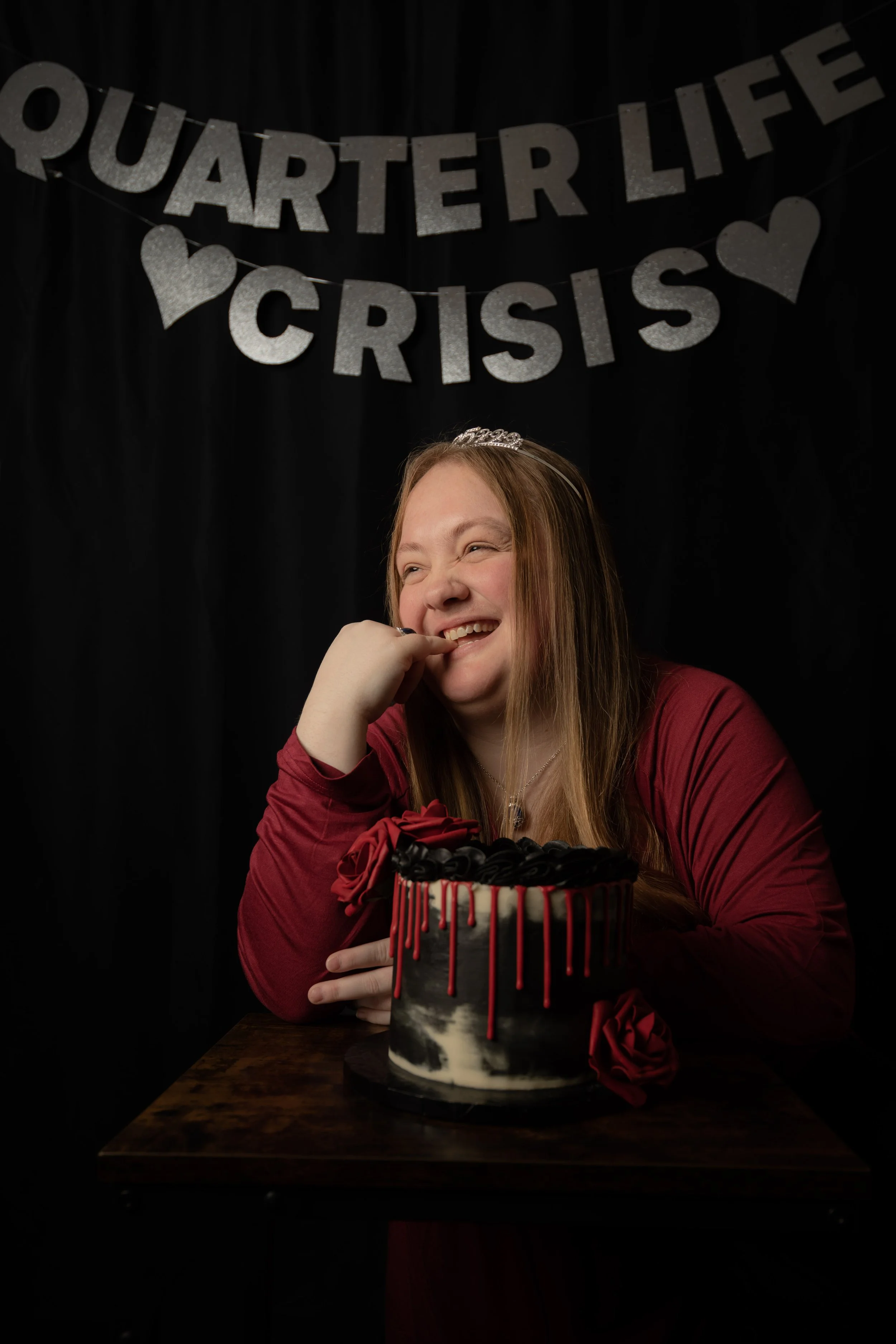A young woman is sitting at a table with a black and white marble cake decorated with red and black roses, smiling and wearing a tiara and a red top. She is in front of a black backdrop with silver glittery words 'Quarter Life Crisis' and hearts hung