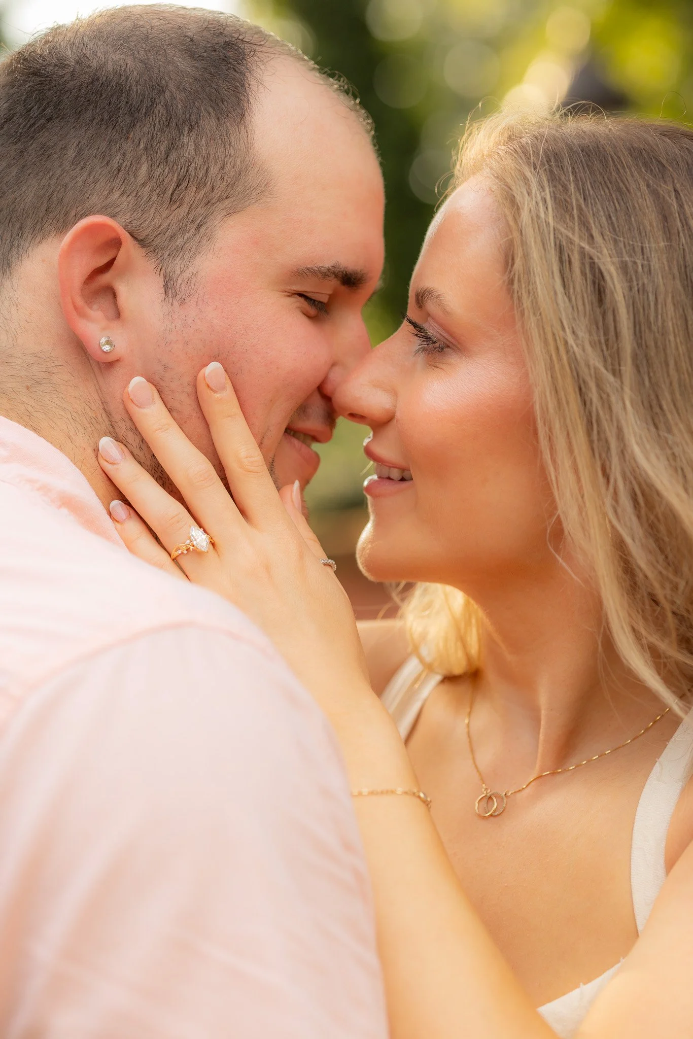 A close-up of a couple with their eyes closed, faces close, smiling, in an outdoor setting with blurred green background, sharing an intimate moment.