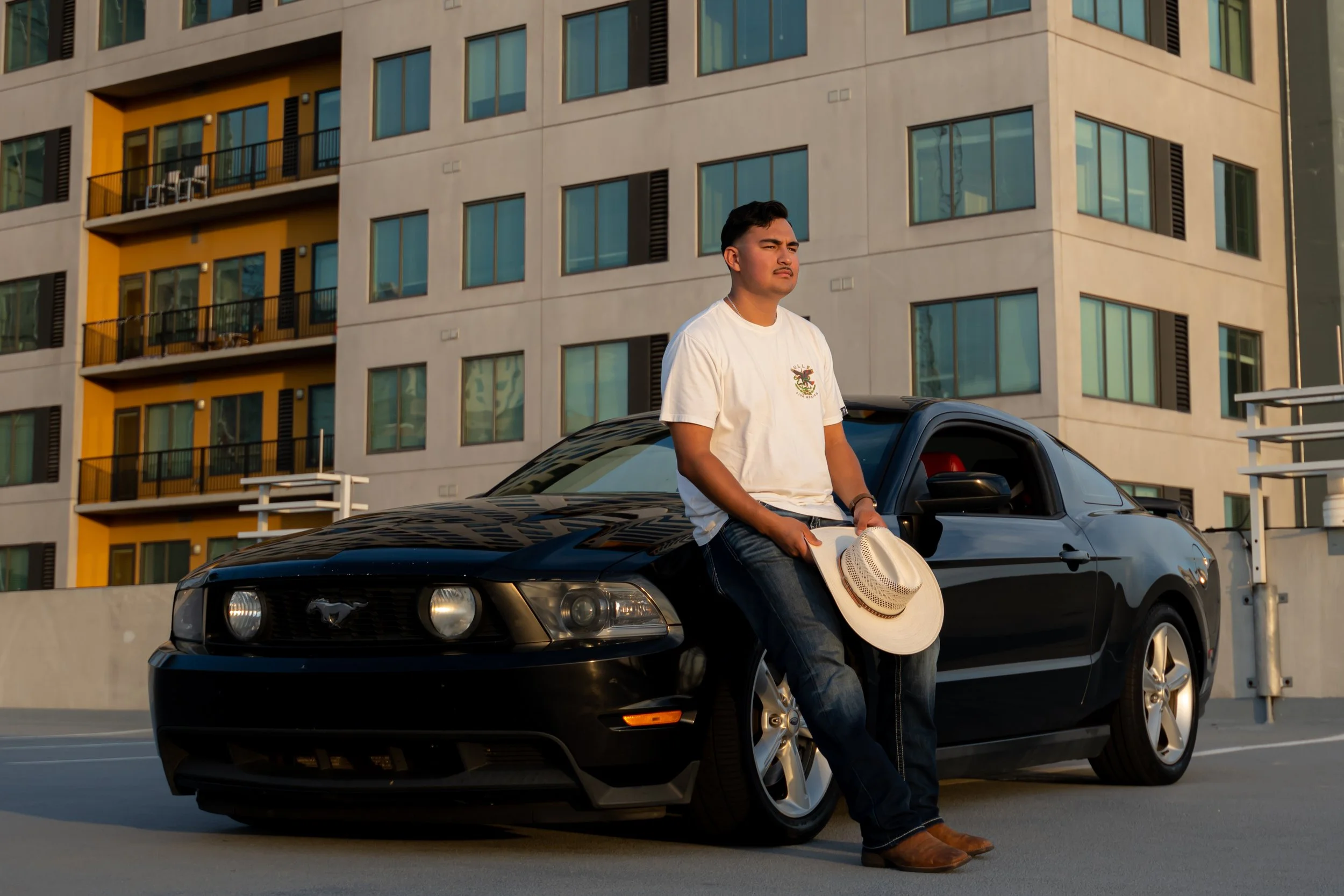 Man in a white T-shirt leaning against a black Ford Mustang parked on a rooftop, holding a straw hat and looking into the distance, with a modern apartment building in the background during sunset.