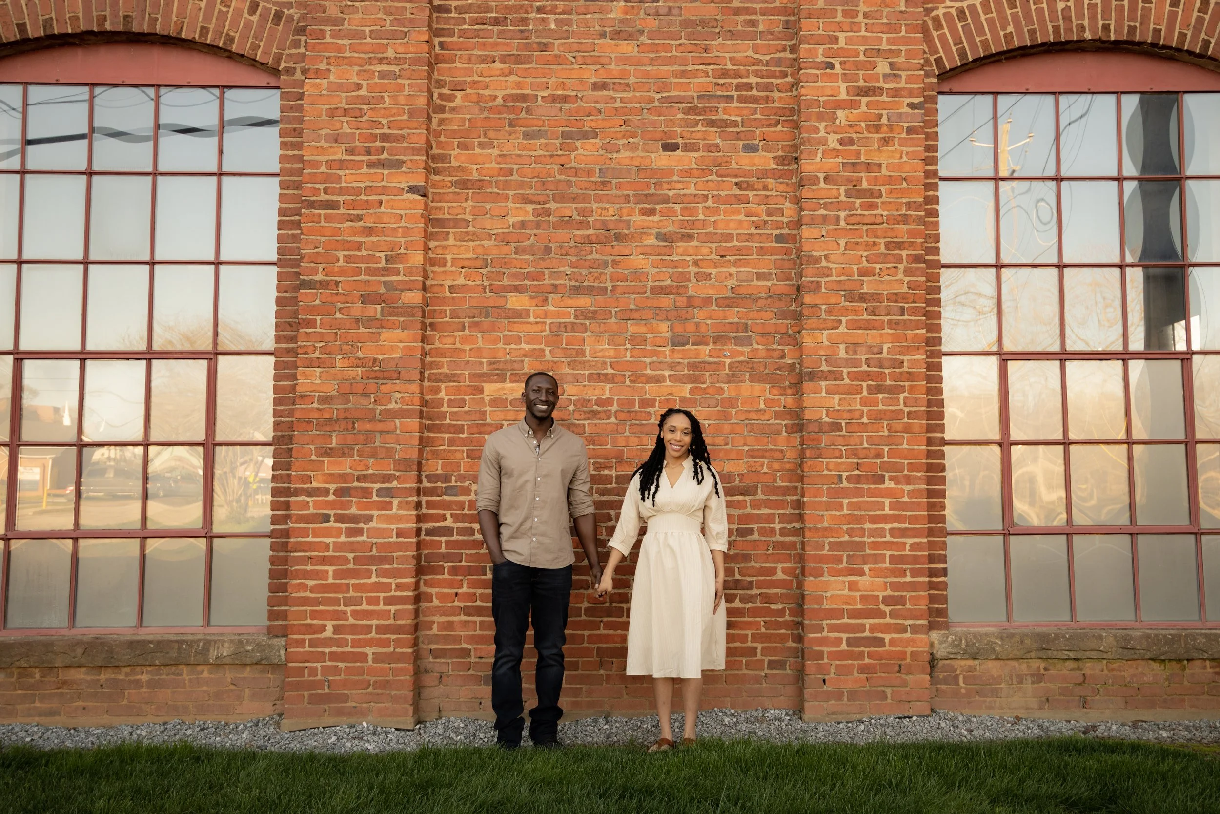 A couple holding hands and smiling in front of a brick building with large windows during sunset.