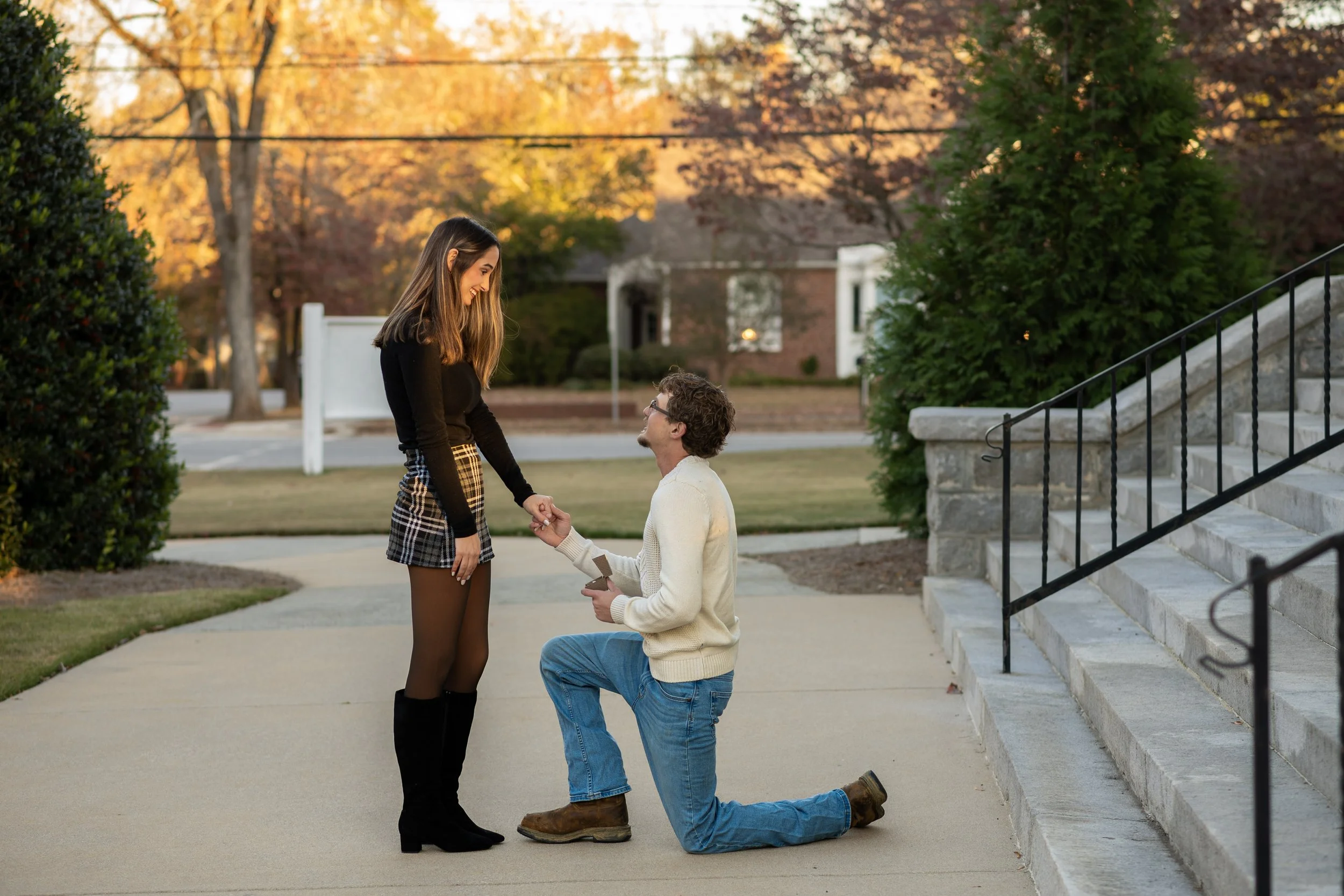 A couple is proposing marriage outdoors during autumn, with the woman standing and the man kneeling on one knee, holding a ring box, while she looks down at him smiling.