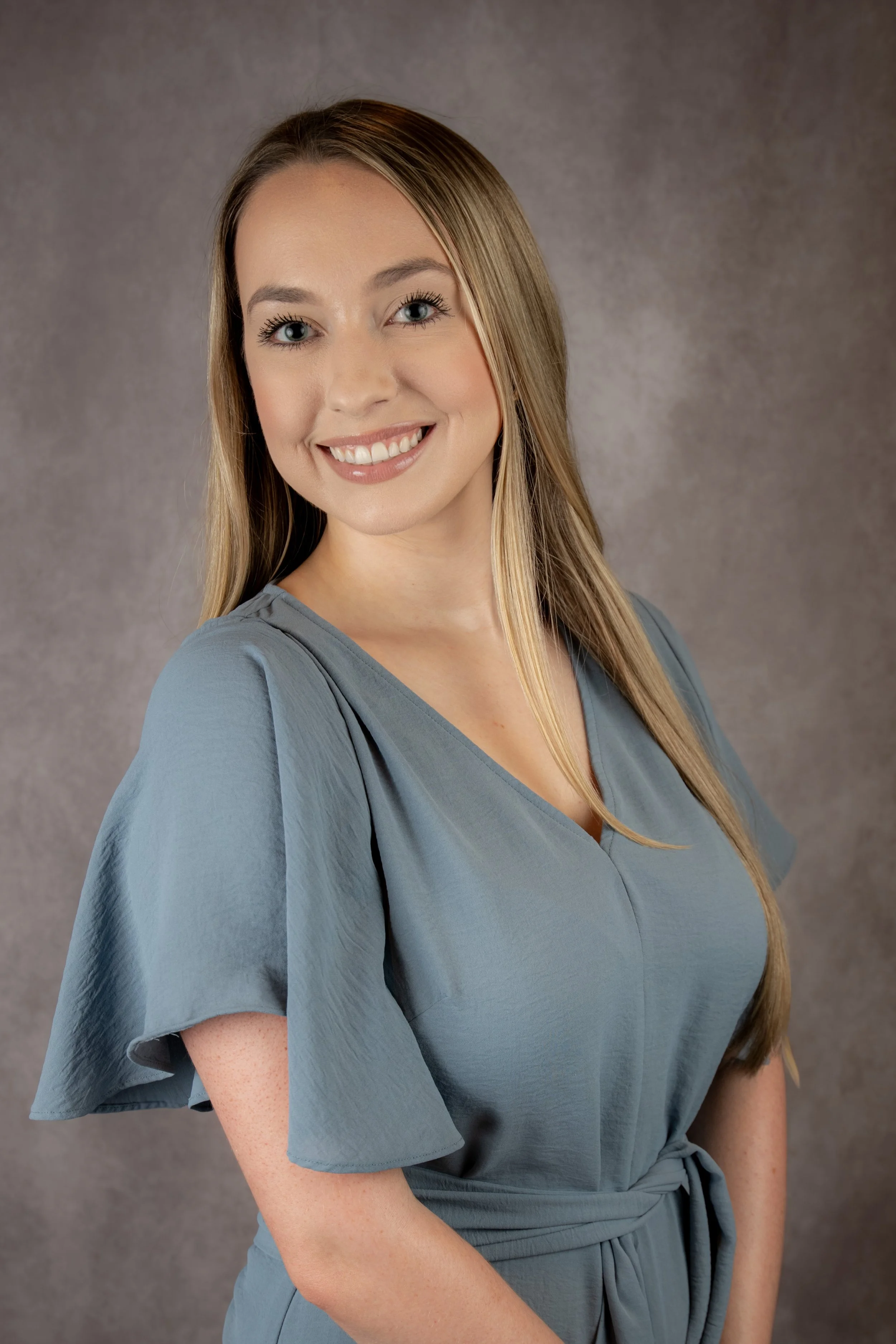 A woman with long blonde hair and blue eyes smiling at the camera, wearing a light blue dress with ruffled sleeves and a waist tie, against a neutral grey background.