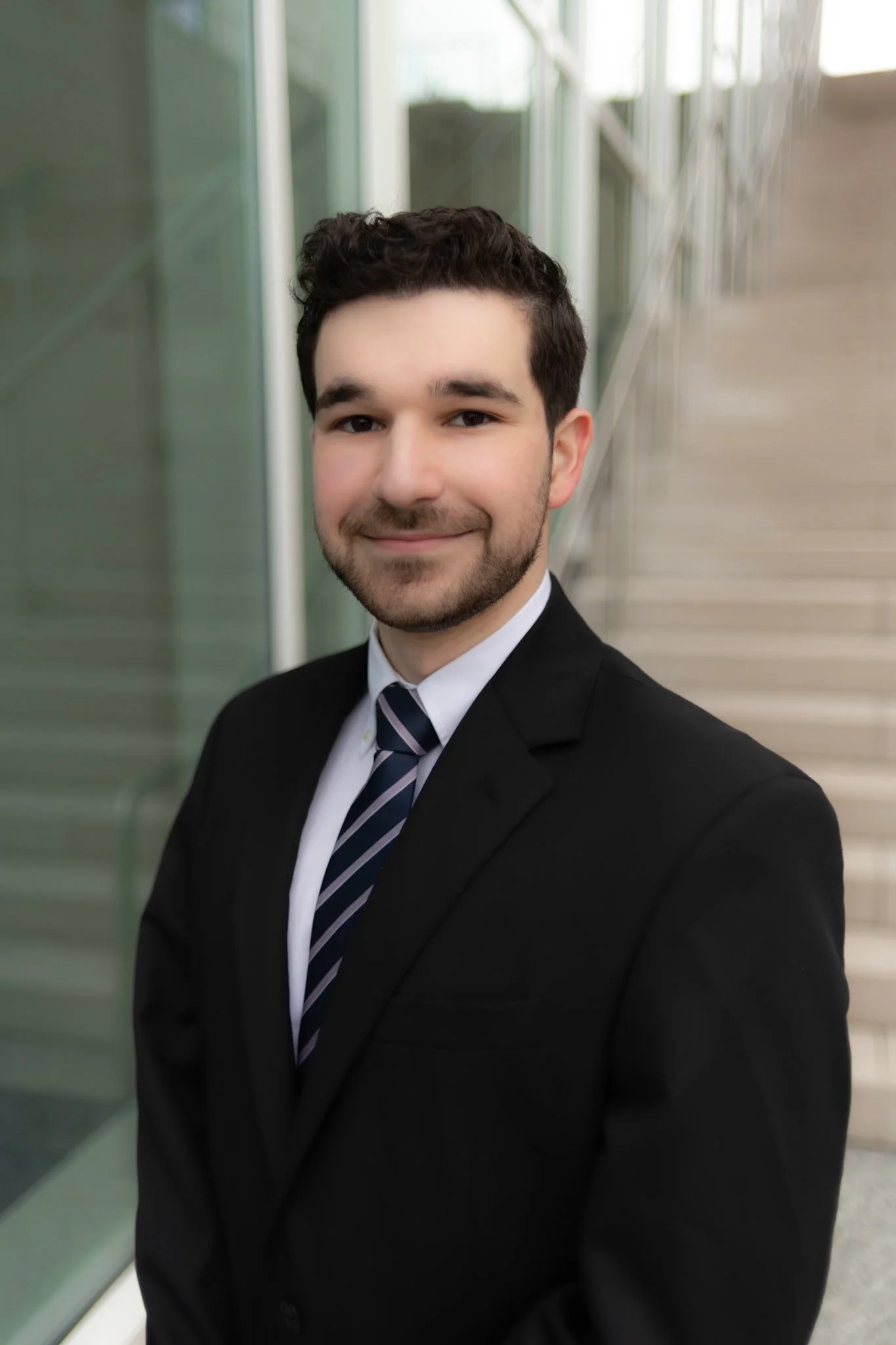 A young man in a black suit, white shirt, and striped tie standing outdoors near a modern building with glass walls and stairs, smiling at the camera.