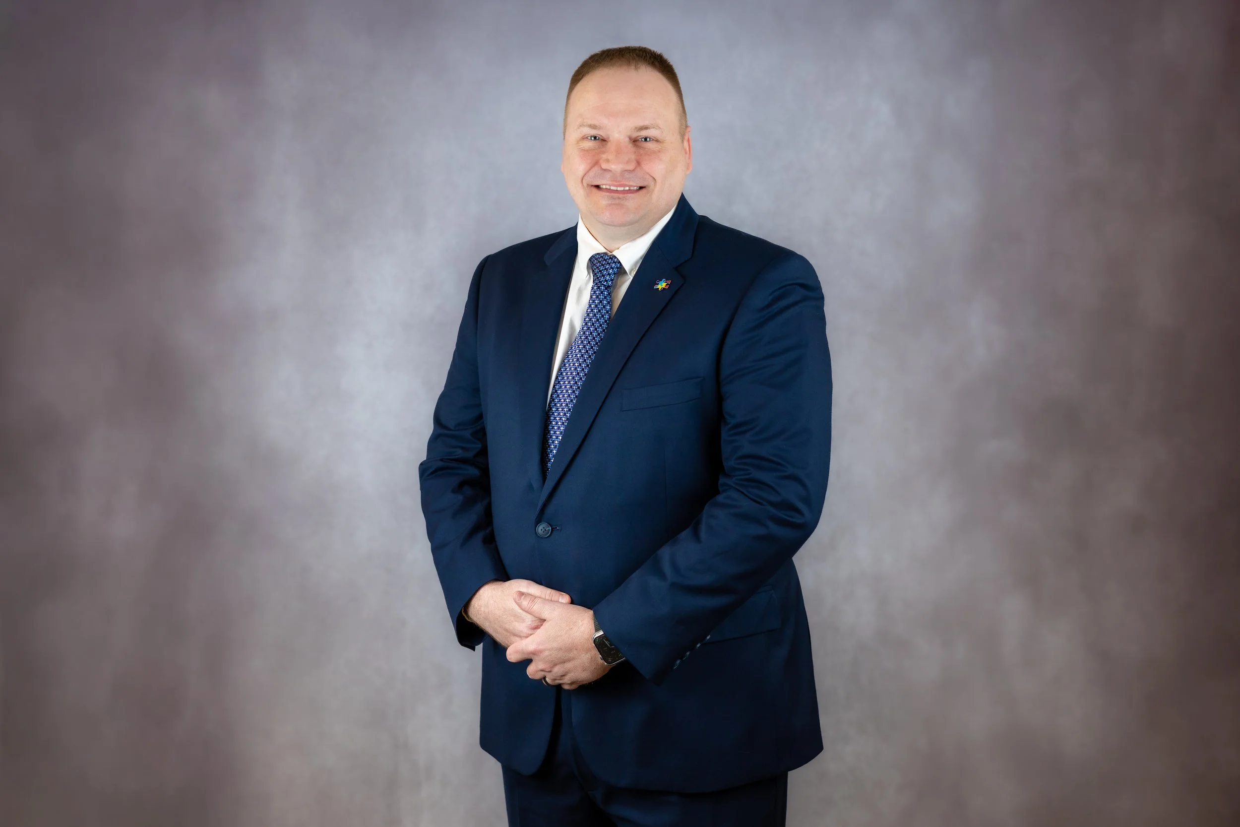 A man in a navy blue suit and patterned tie standing against a plain gray background, smiling at the camera.