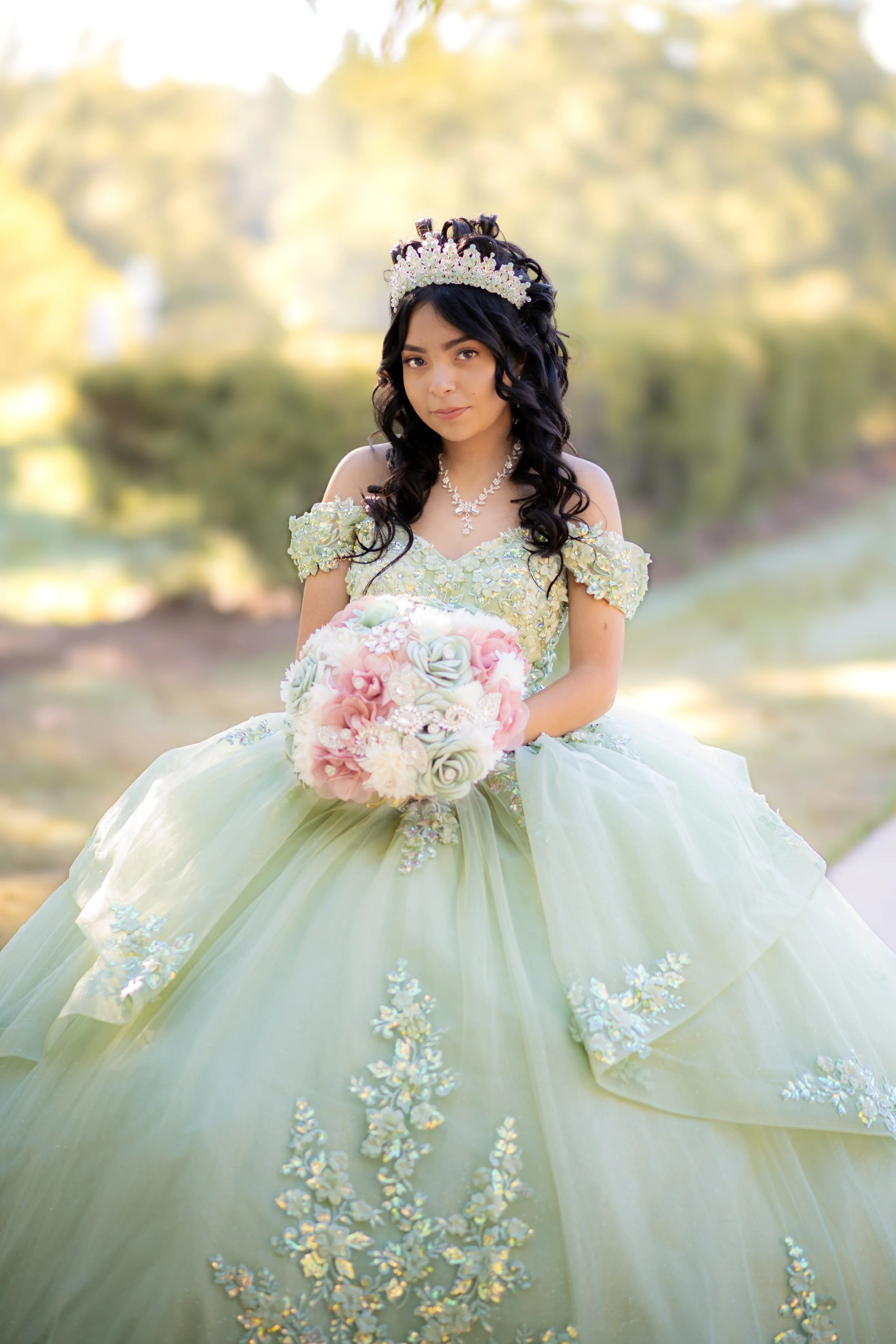 A young girl in a pastel green ball gown with floral embellishments holds a bouquet of pink and white flowers while standing outdoors with a blurred natural background, wearing a tiara and jewelry.