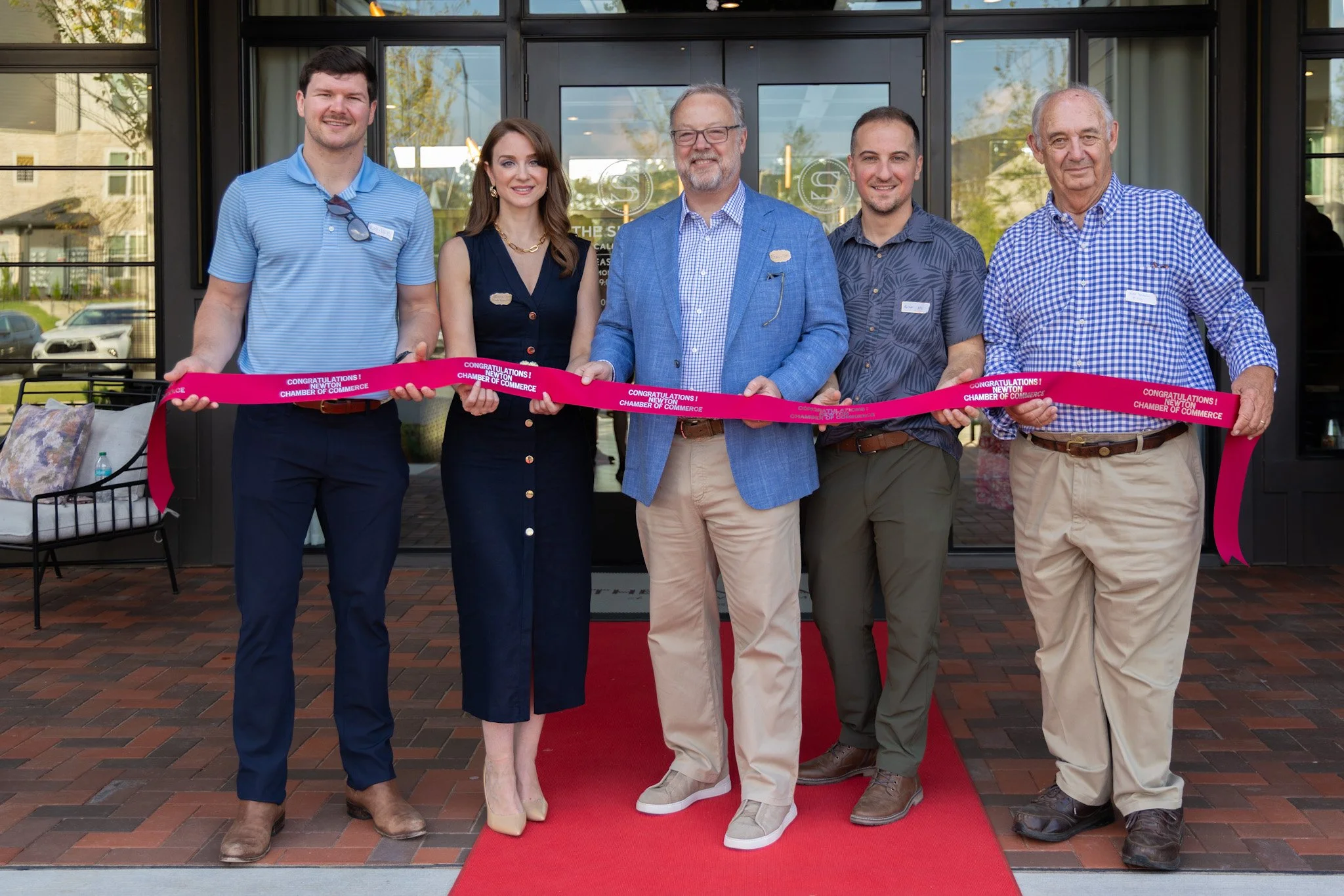 Six people standing outside a building, holding a pink ribbon for a ribbon-cutting ceremony, with a red carpet.