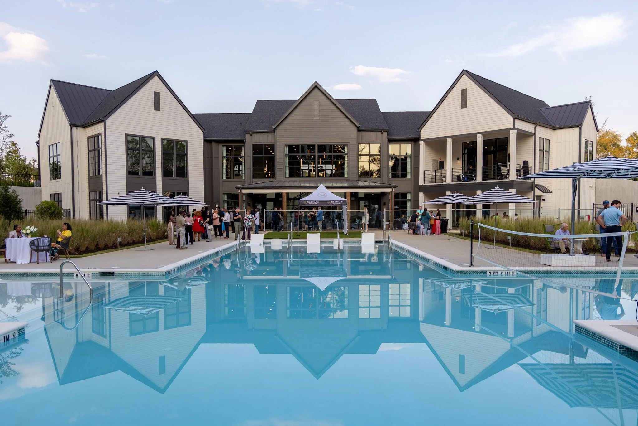 A poolside scene at a residential complex with a large, modern building in the background. There are people socializing, some standing and some seated under umbrellas, near the pool and on the balconies.