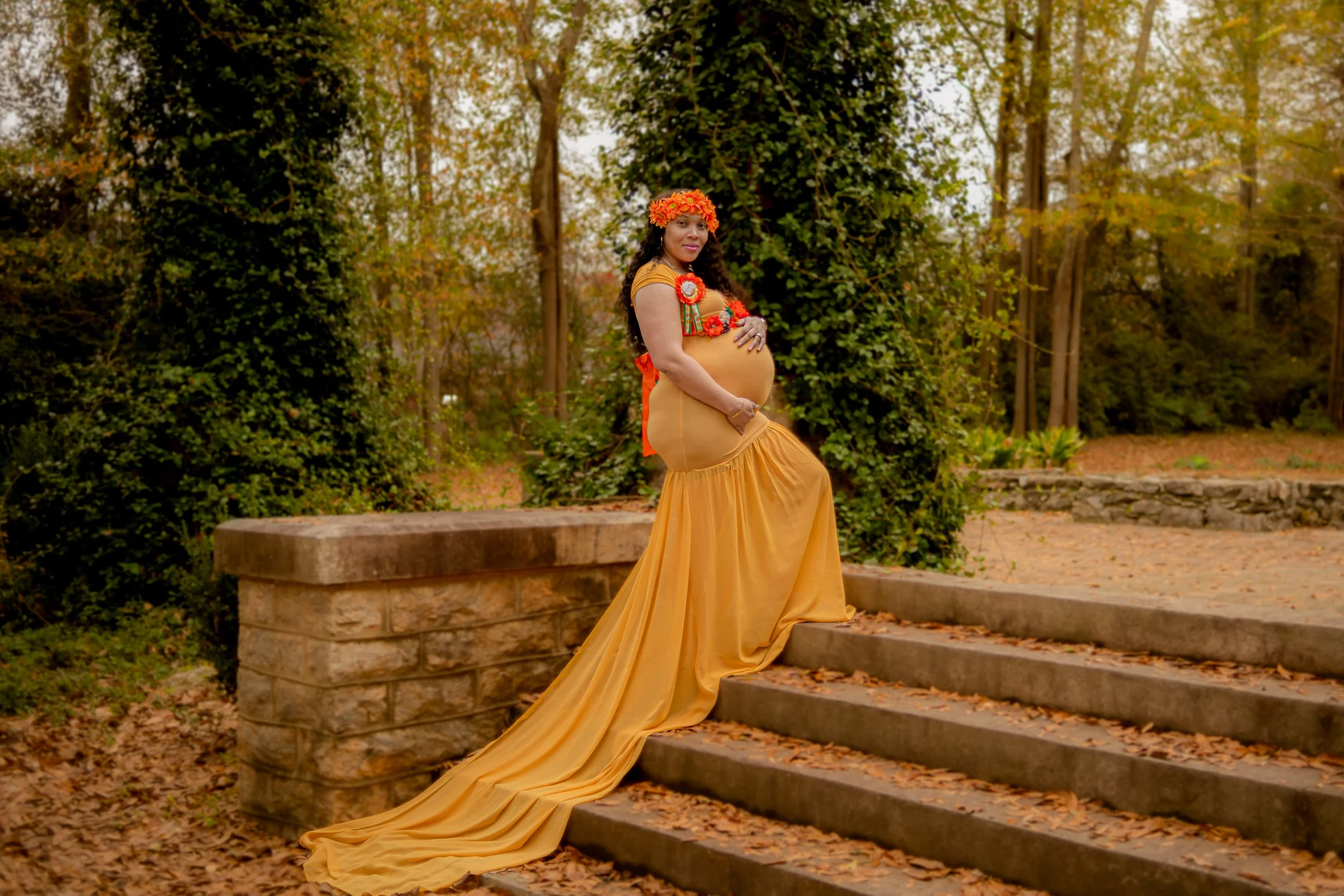 A pregnant woman in a gold-colored dress with a long train, standing outdoors on fallen autumn leaves, surrounded by trees and greenery, wearing a floral crown and flower decorations.