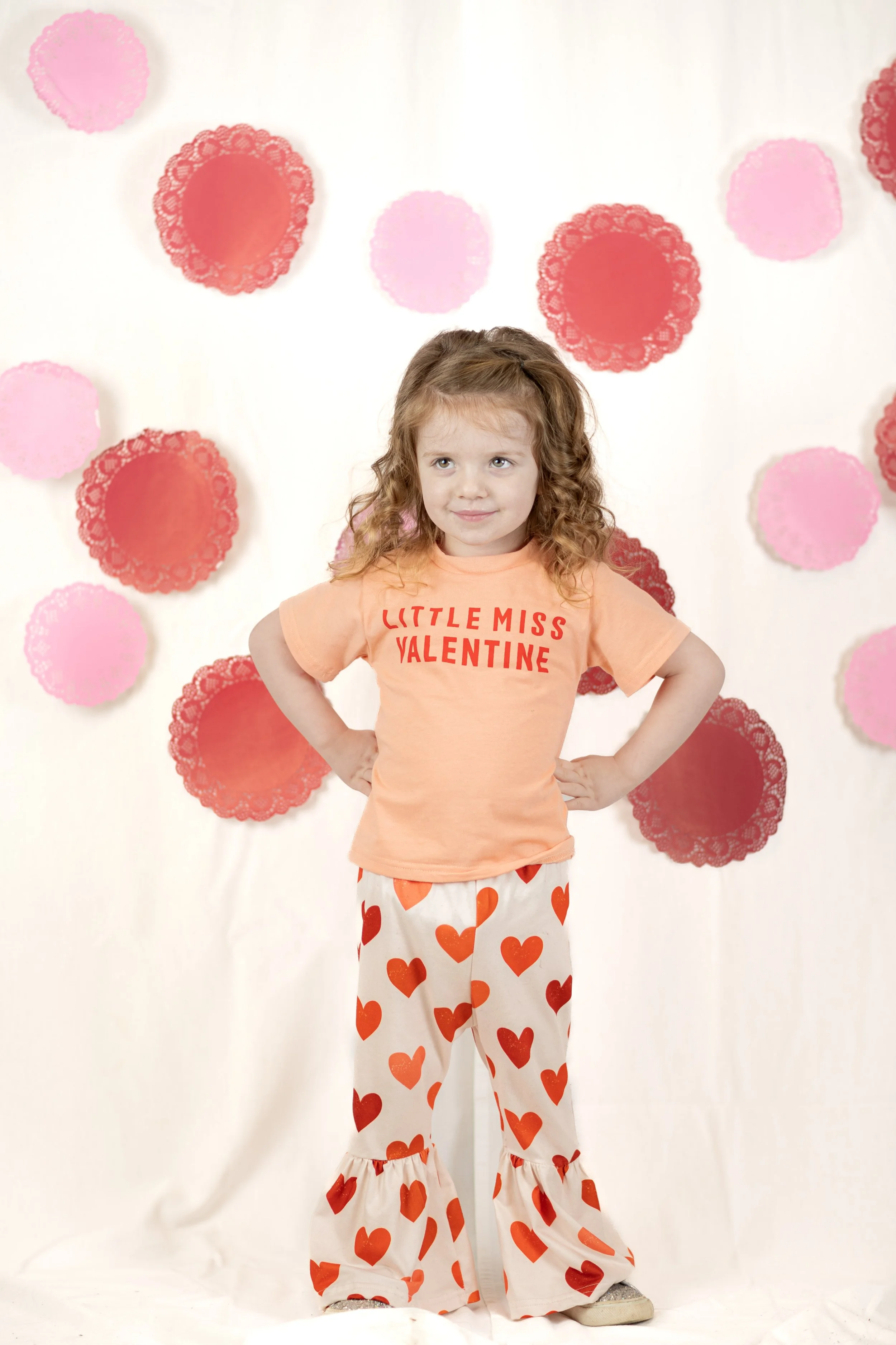 A young girl with curly red hair stands with her hands on her hips in front of a backdrop decorated with pink and red paper doilies. She is wearing a peach t-shirt that says 'LITTLE MISS VALENTINE' and white pants with red and orange hearts.