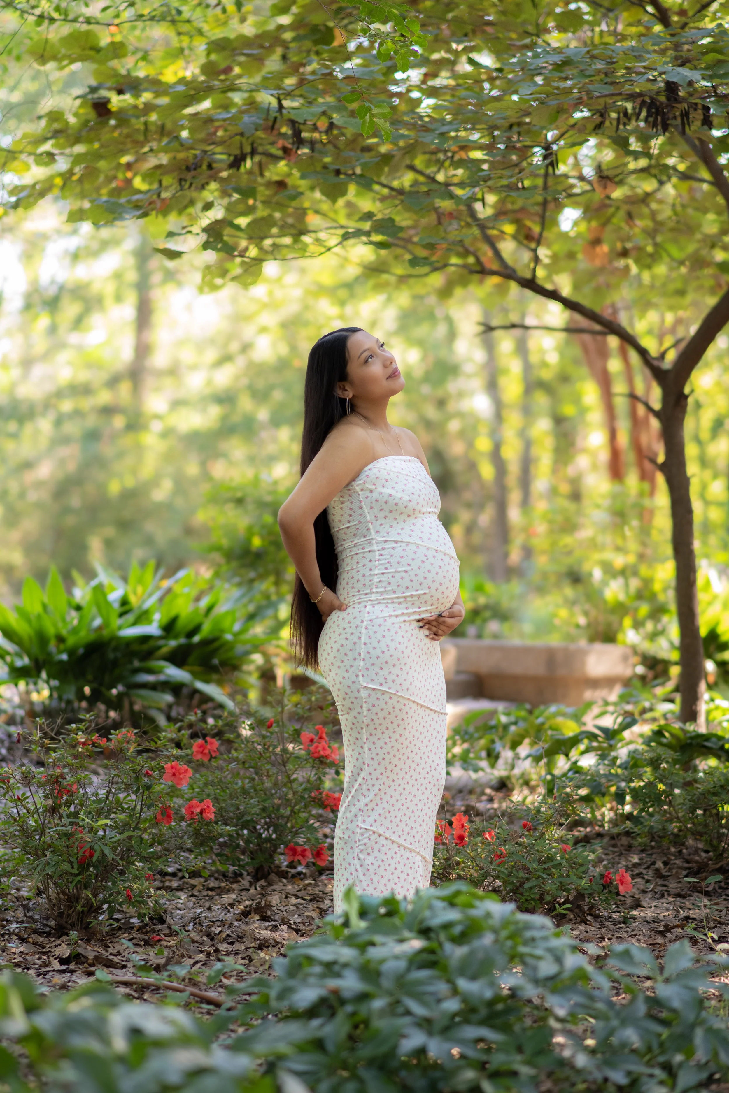 Pregnant woman in a white dress with pink flowers standing in a lush garden, gently holding her belly and gazing upward among green trees and plants.