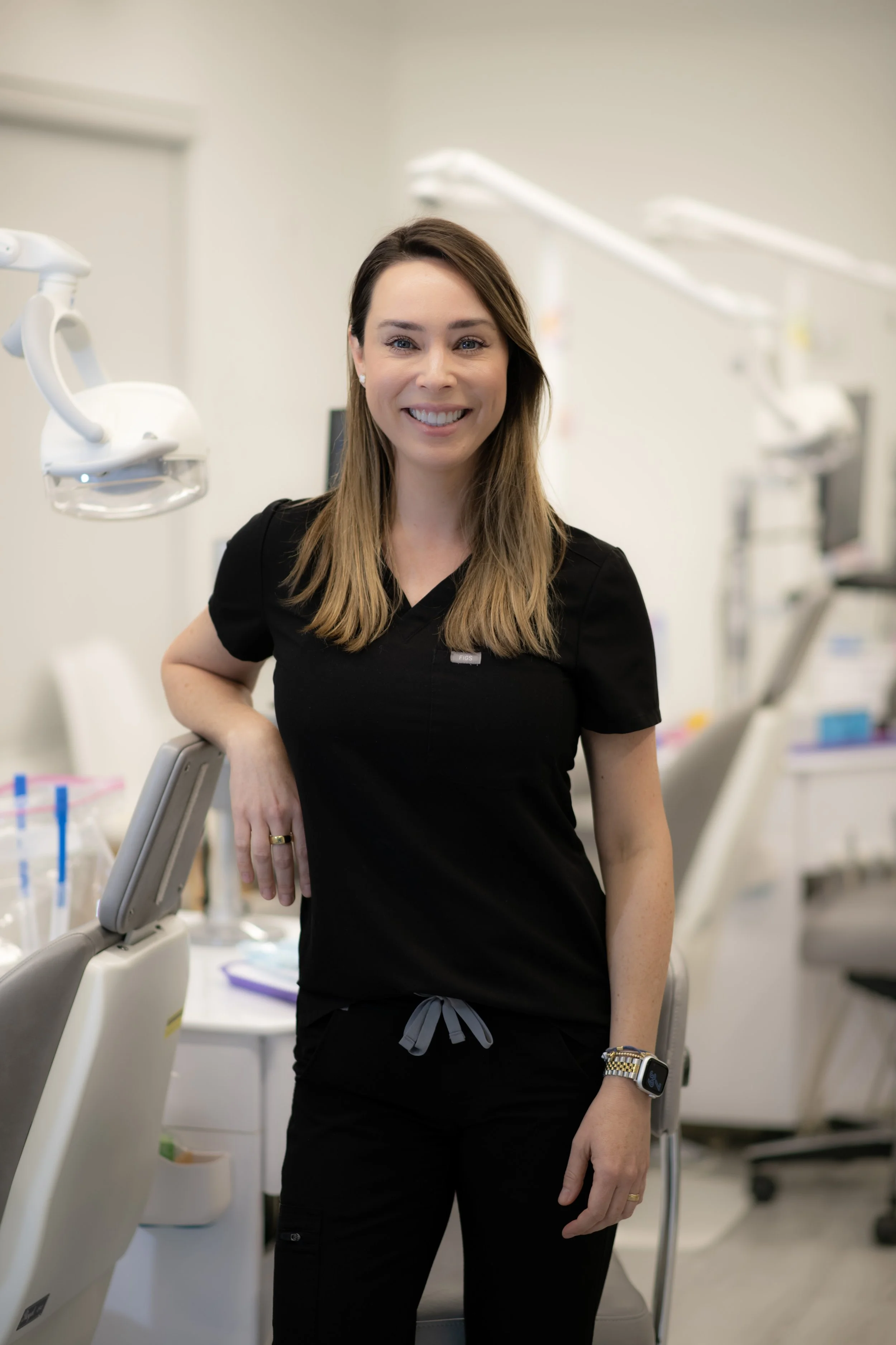 A female healthcare professional in black scrubs standing in a medical office or clinic, smiling at the camera with dental equipment in the background.