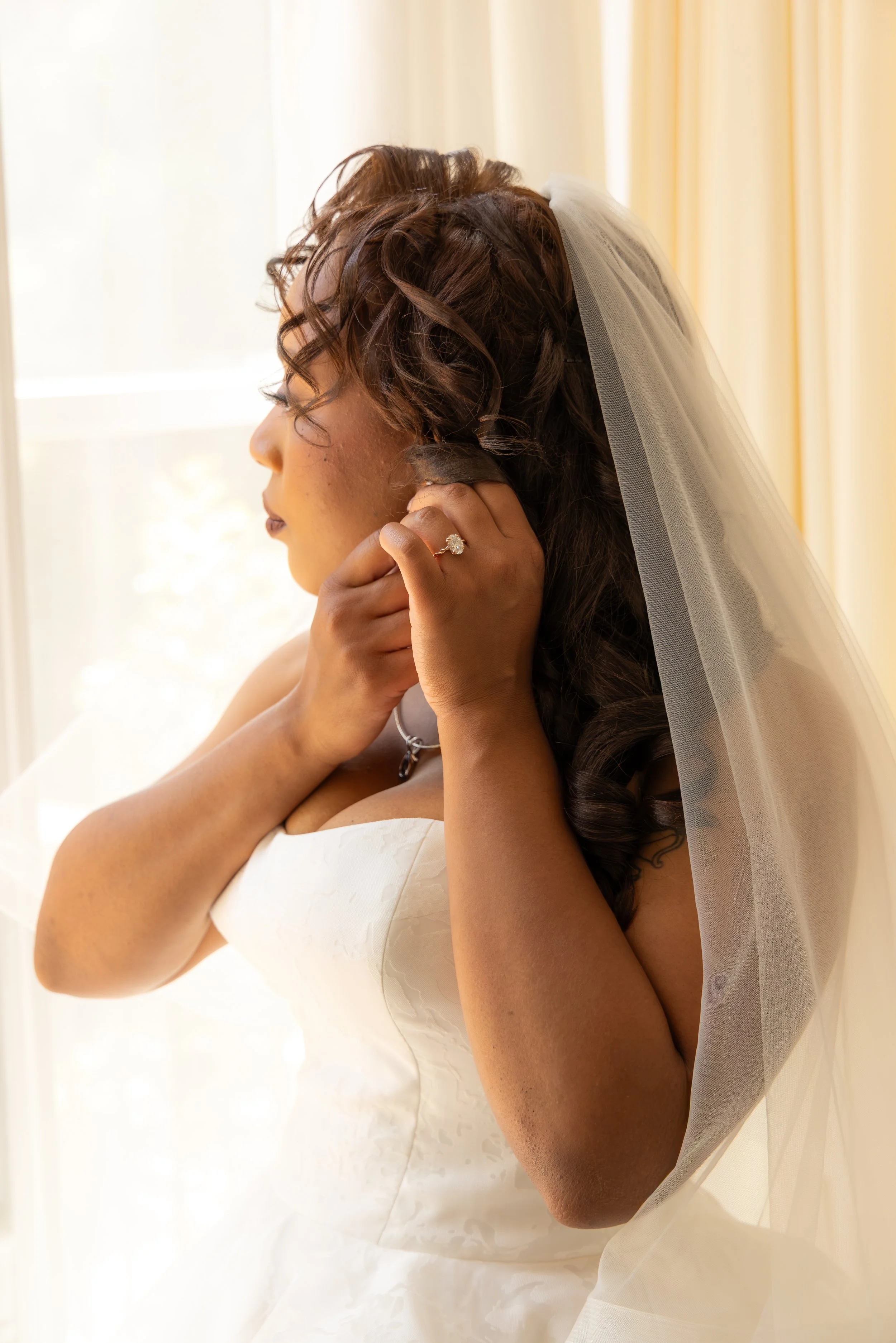 A bride with curly brown hair wearing a white wedding dress and veil, holding an earring to her ear.