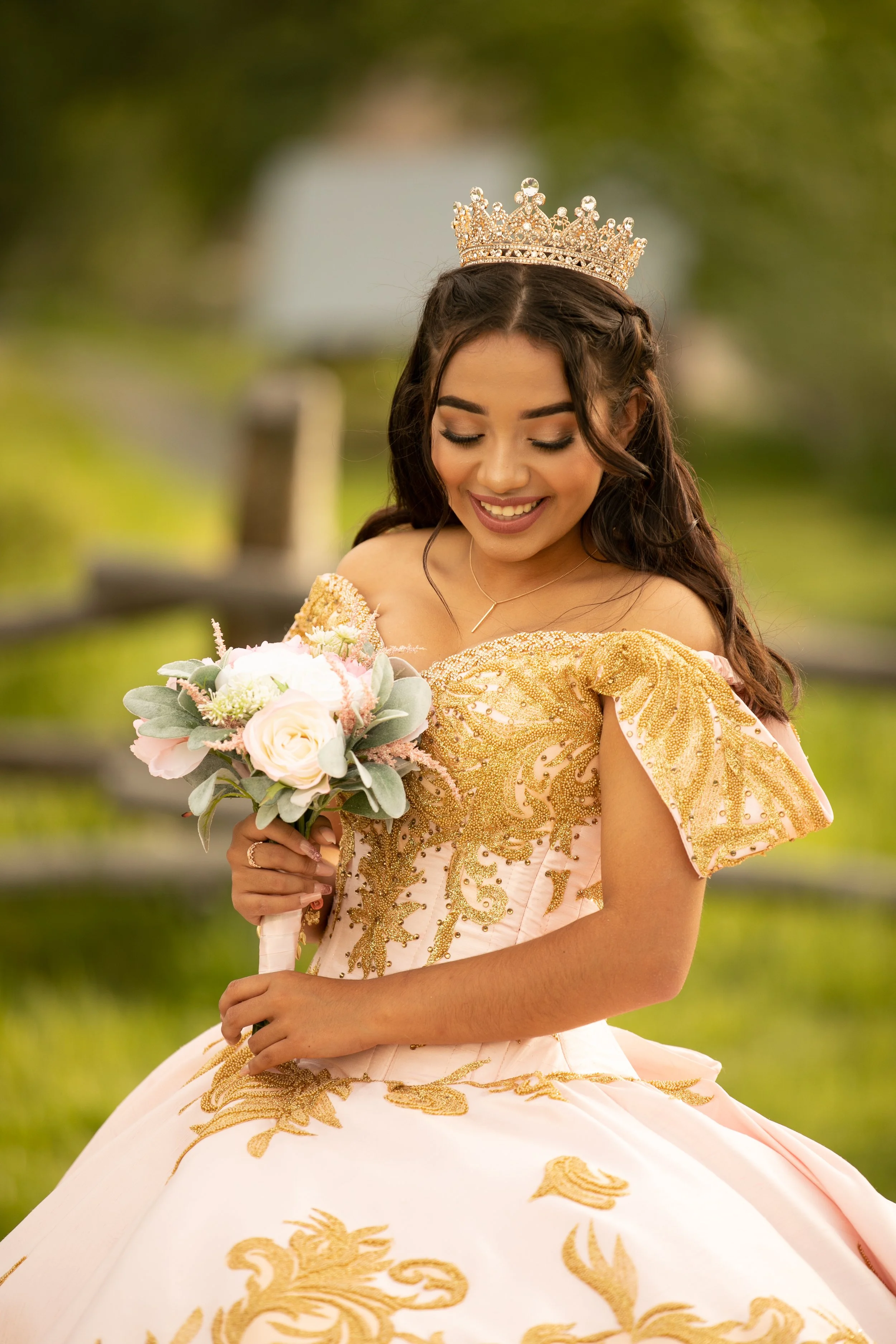 A young woman dressed in a pink and gold gown with ornate embroidery, wearing a crown, and holding a bouquet of flowers, standing outdoors with green foliage in the background.
