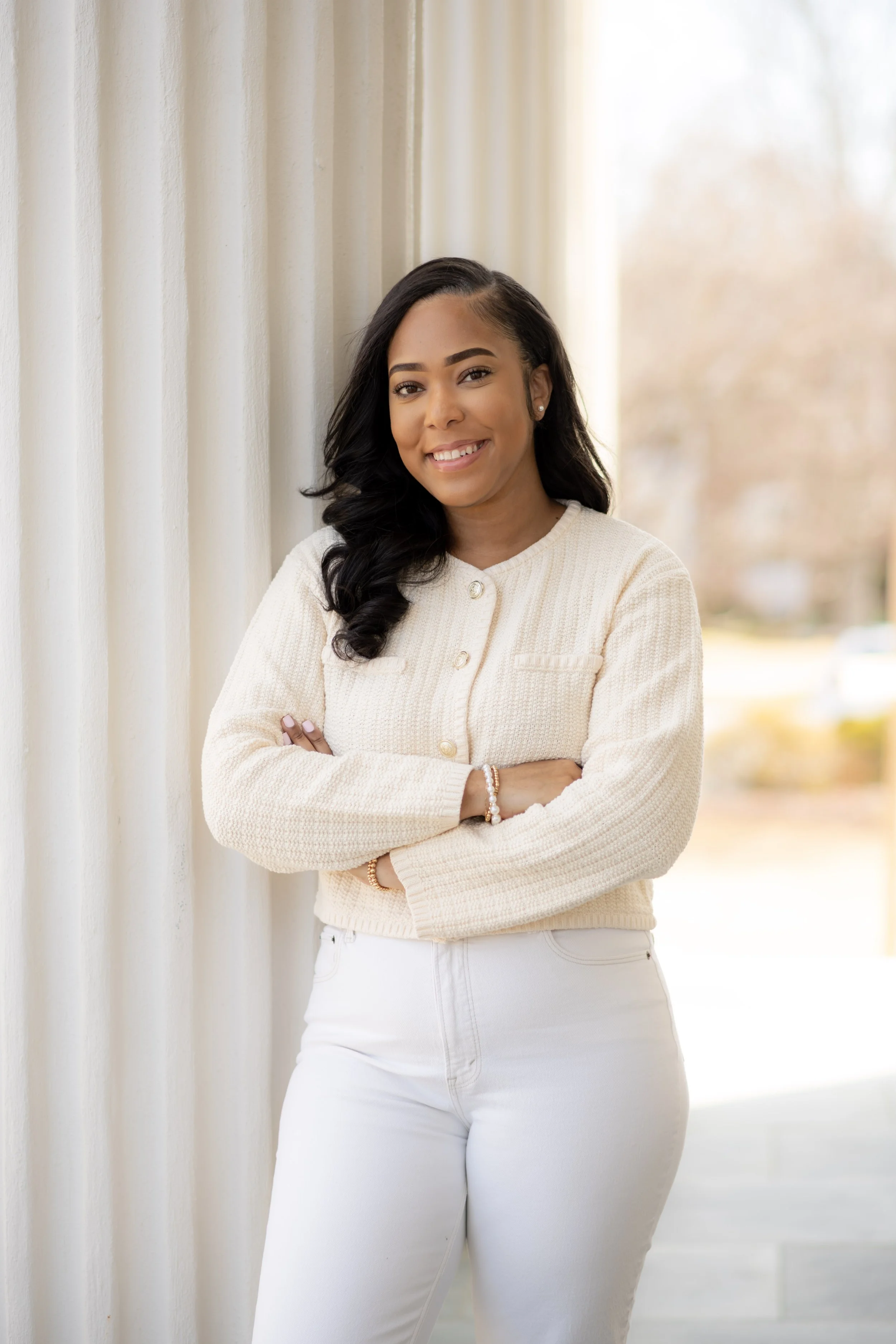 Young African American woman with black hair, smiling, crossing arms, standing next to cream curtains outdoors.