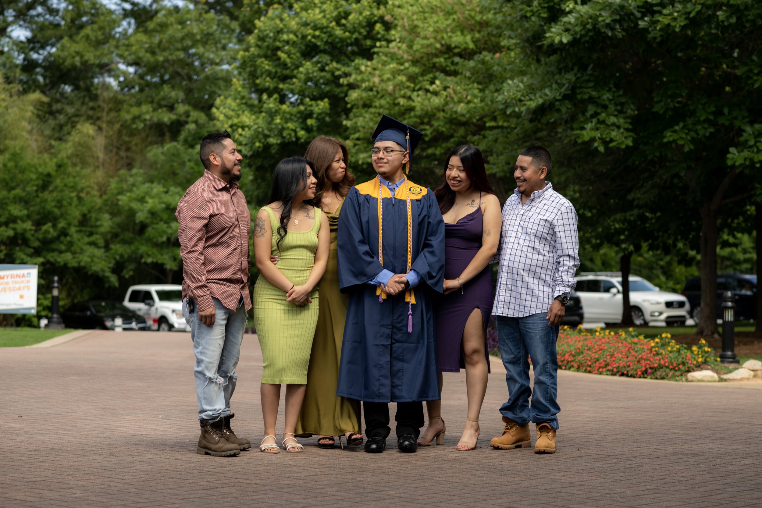 A group of six people, including a man in a blue graduation gown and cap, pose outdoors on a paved path with greenery in the background. The graduate is at the center, surrounded by friends or family members.