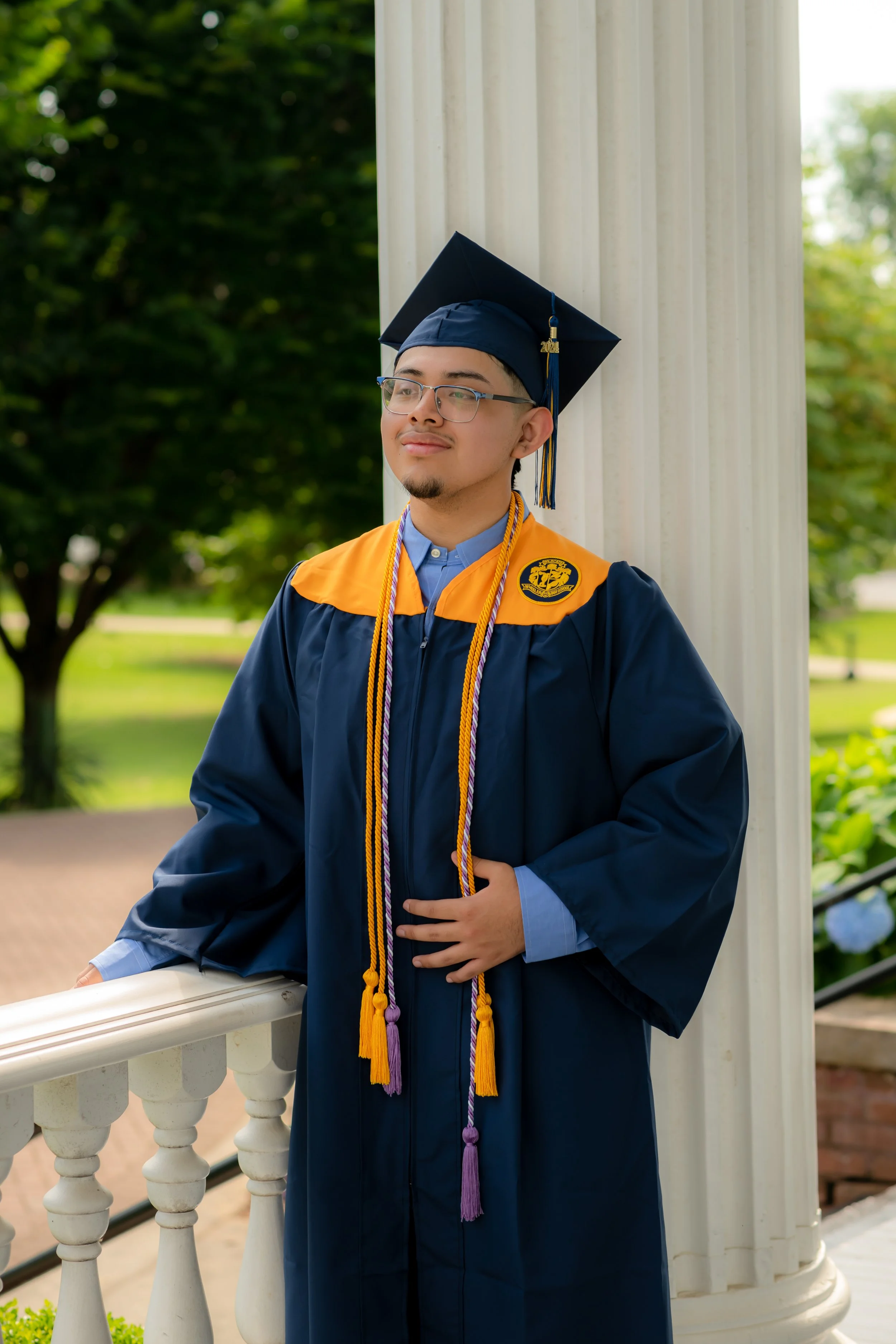 Young man in a graduation cap and gown, standing outdoors by a white column, with a green park in the background.