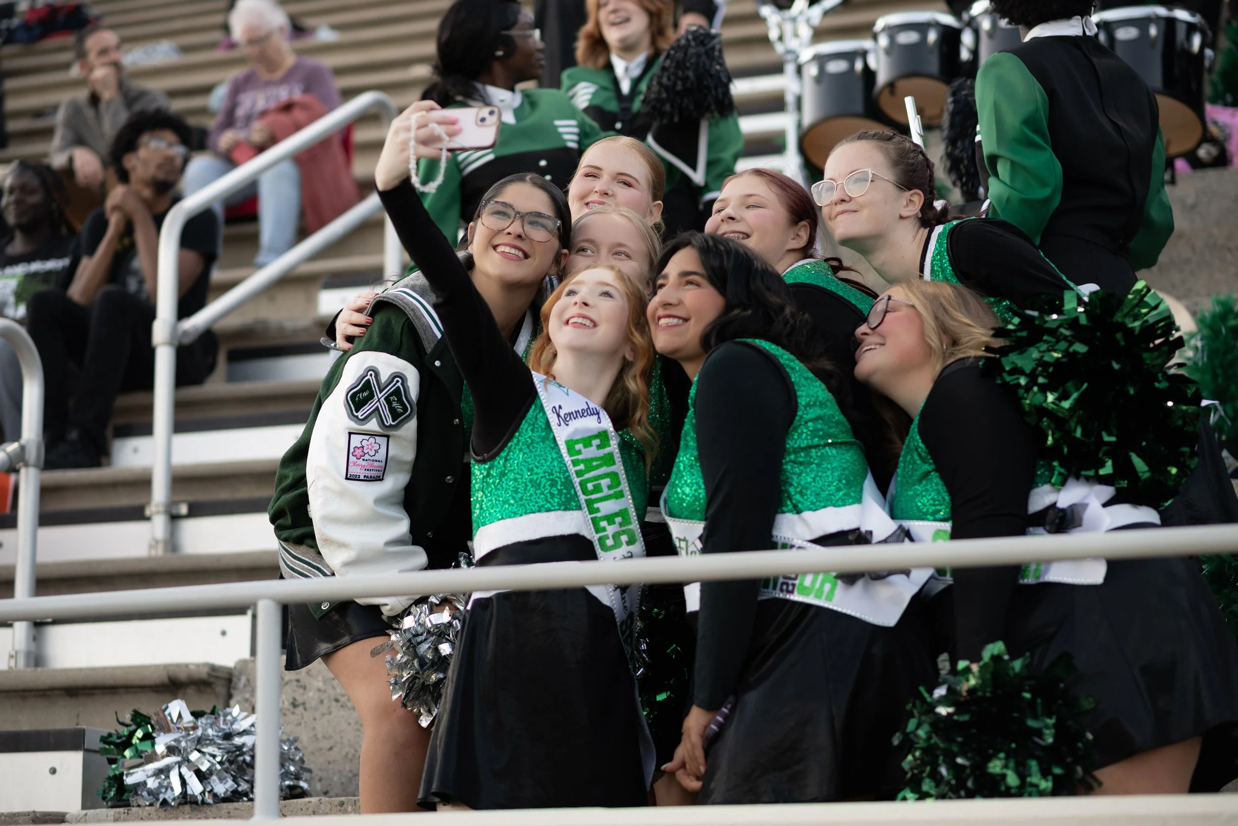 Group of cheerleaders in green and black uniforms taking a selfie at a sports event, with spectators in the stands behind them.