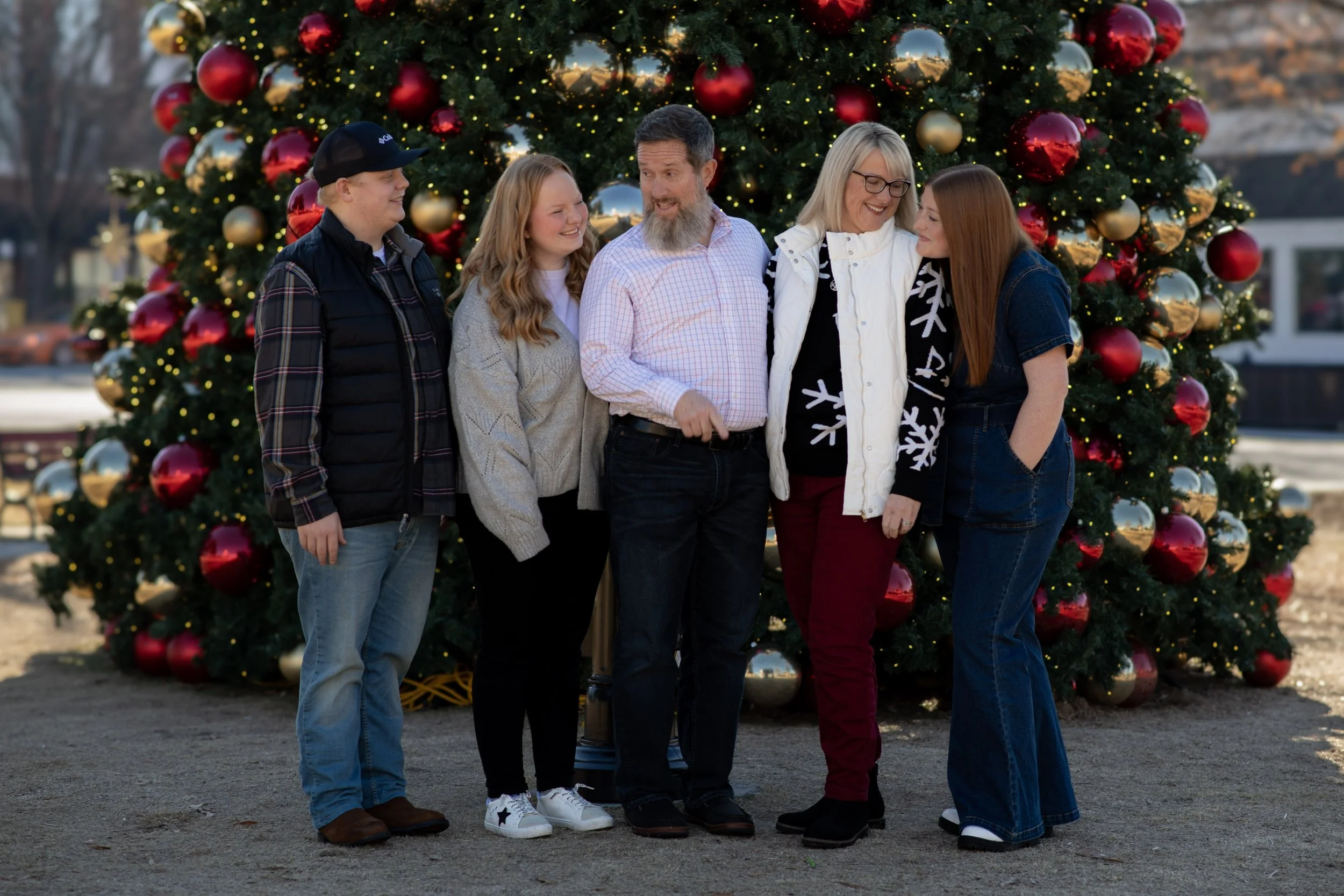 A family of five standing outdoors in front of a decorated Christmas tree, smiling and enjoying the holiday season.