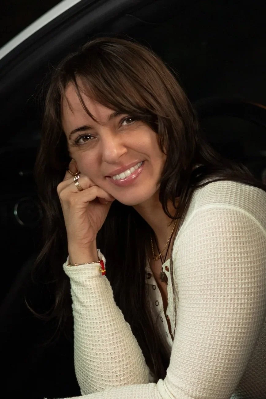 A woman with brown hair and a white textured long-sleeve top, smiling and resting her chin on her hand, inside a car.