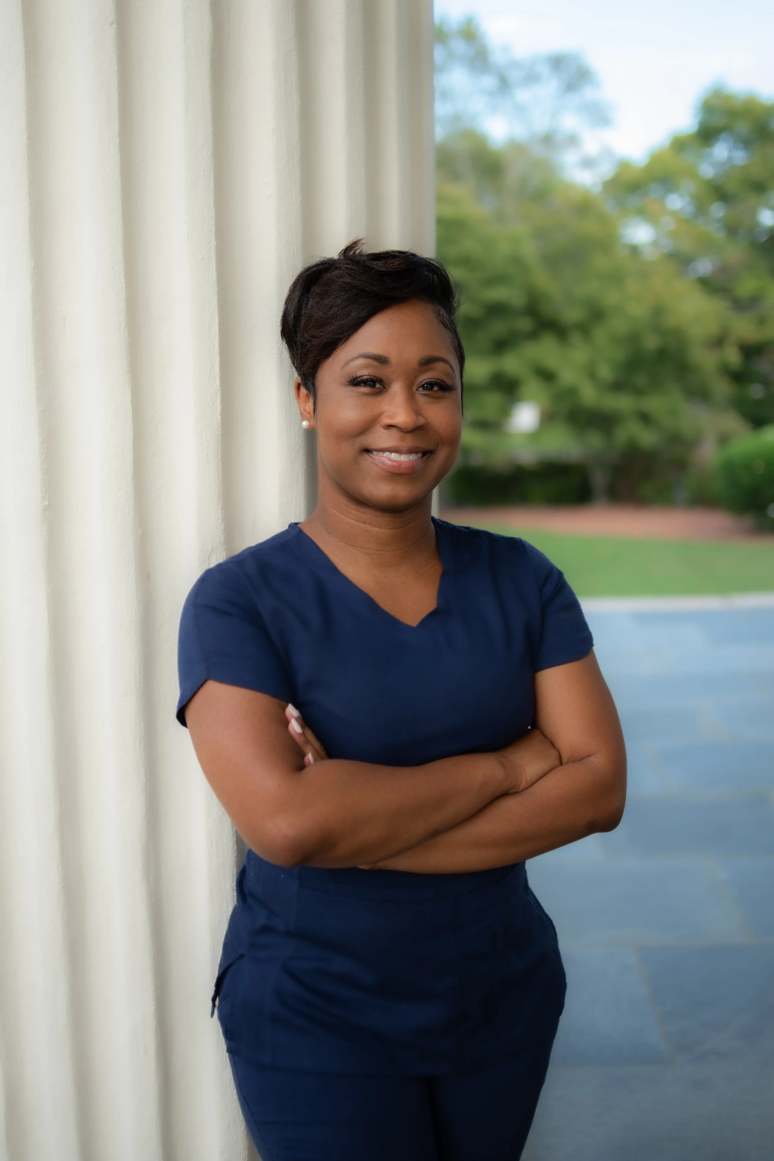 A woman with short dark hair, wearing a navy blue uniform, standing outdoors near a white column with green trees and a blue sky in the background, smiling with arms crossed.
