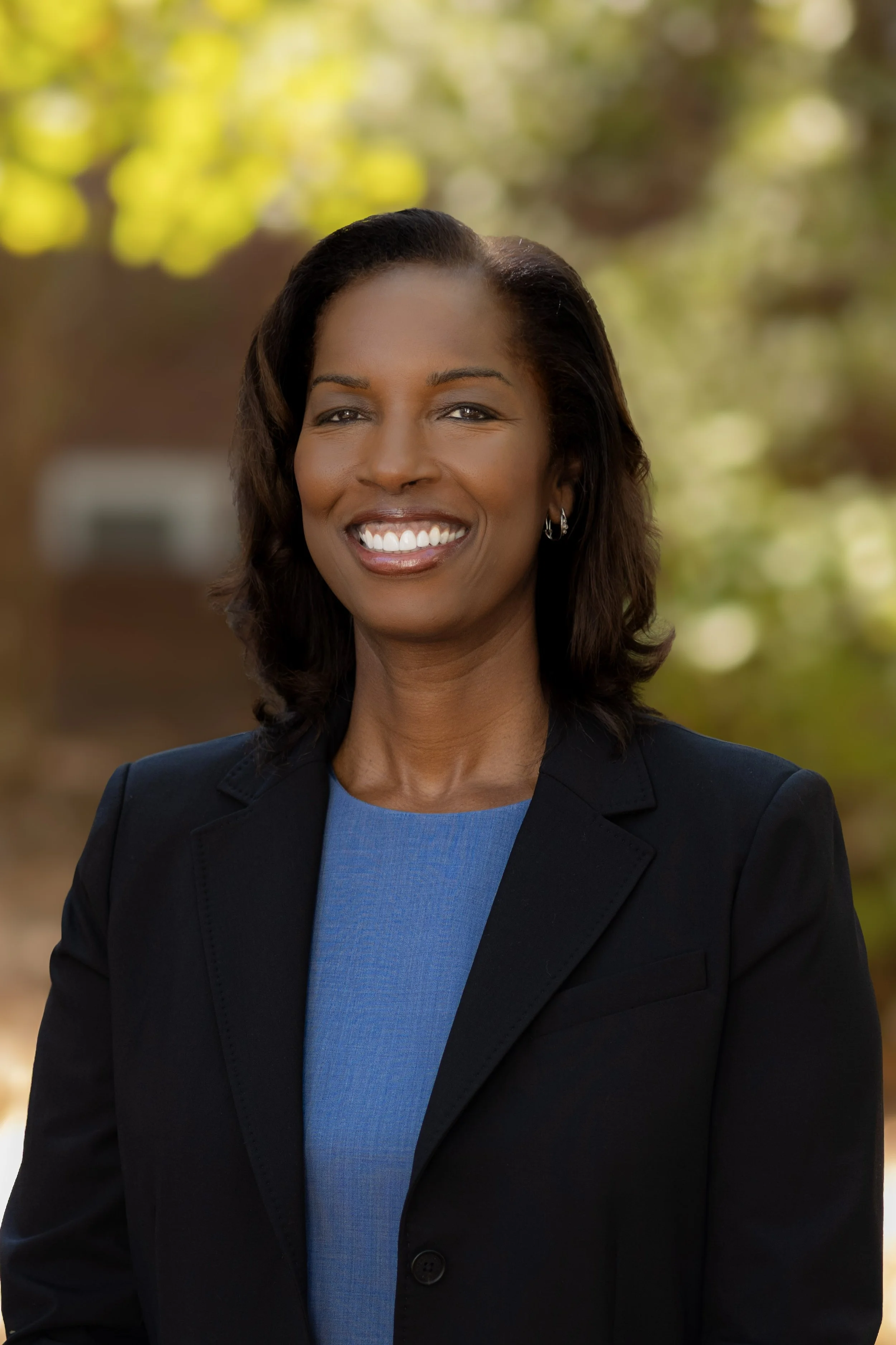 A professional woman smiling outdoors, wearing a black blazer and blue top, with a blurred background of green and yellow foliage.