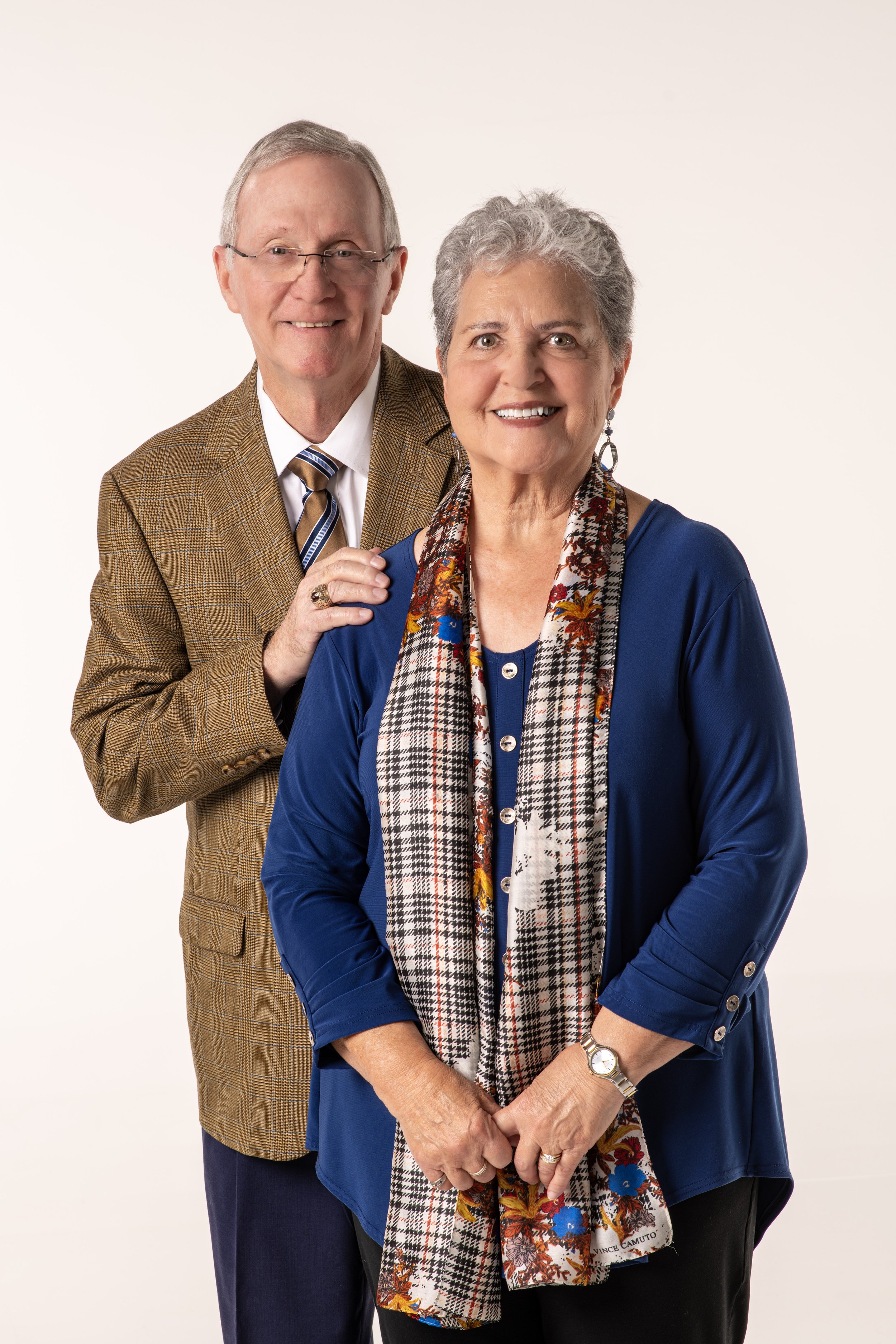 A smiling elderly couple, man in a brown plaid jacket and woman in a blue cardigan with a colorful scarf, standing against a white background.