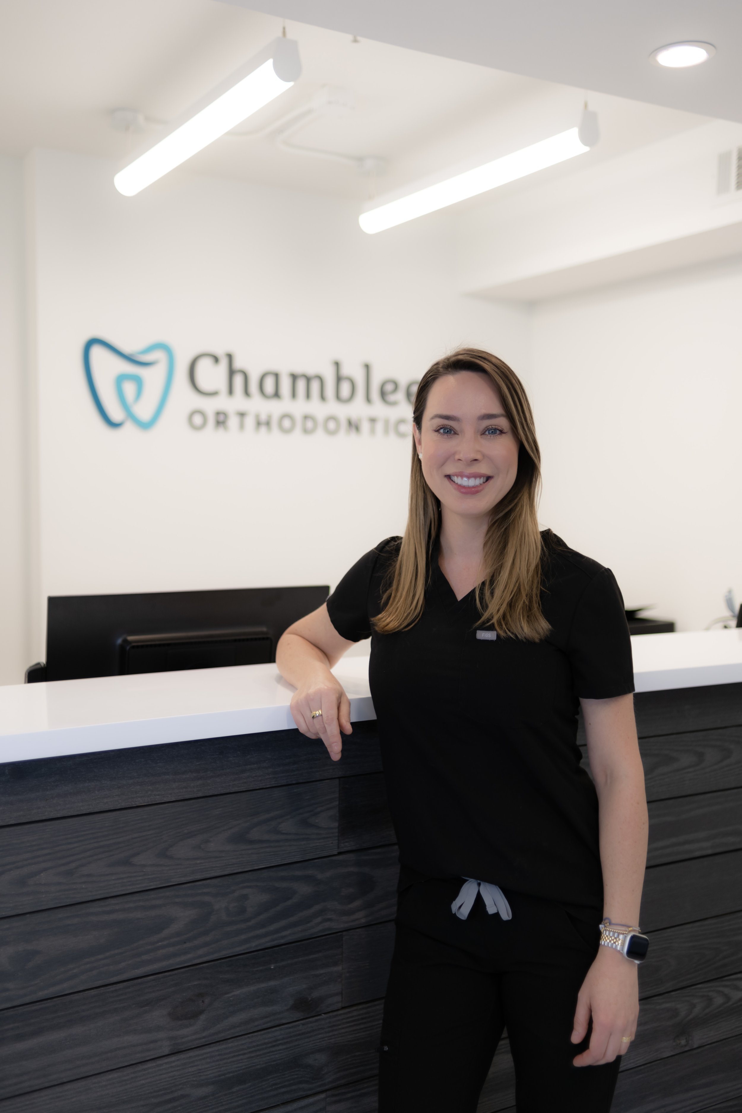A woman, Dr. C, in black scrubs leaning on a reception desk at Chamblee Orthodontics, smiling at the camera.