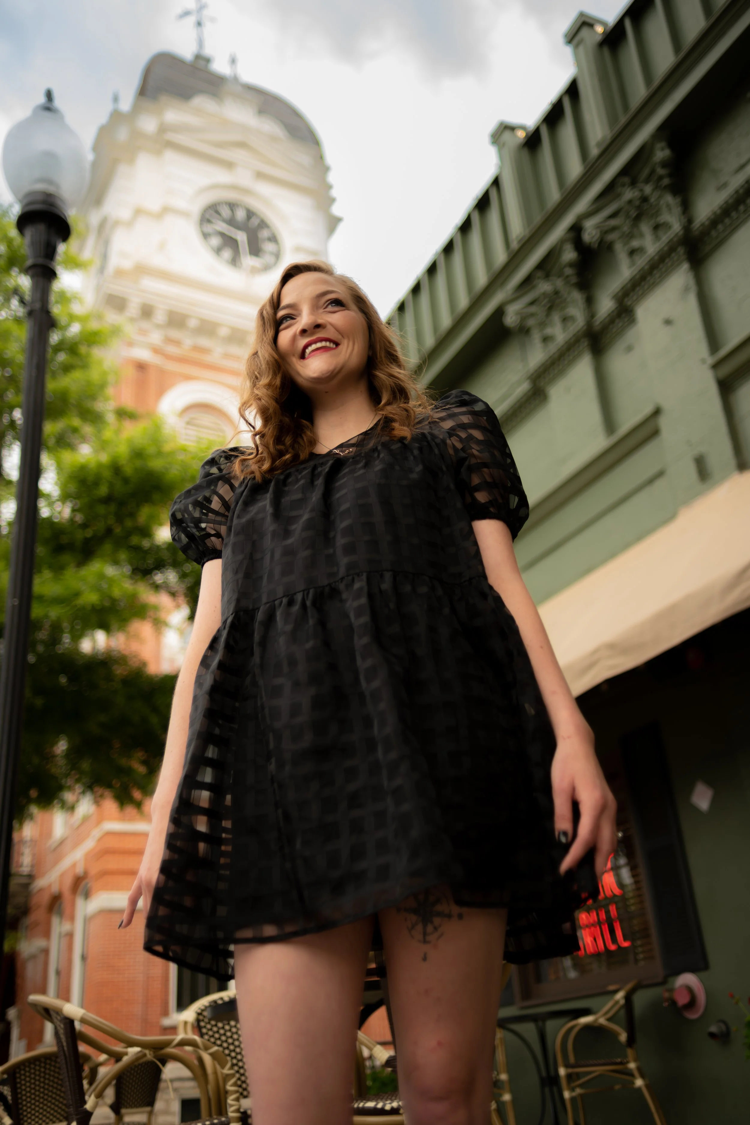 A woman with curly hair smiling and standing outdoors in front of a historic building with a clock tower, in an urban area with greenery and outdoor seating.