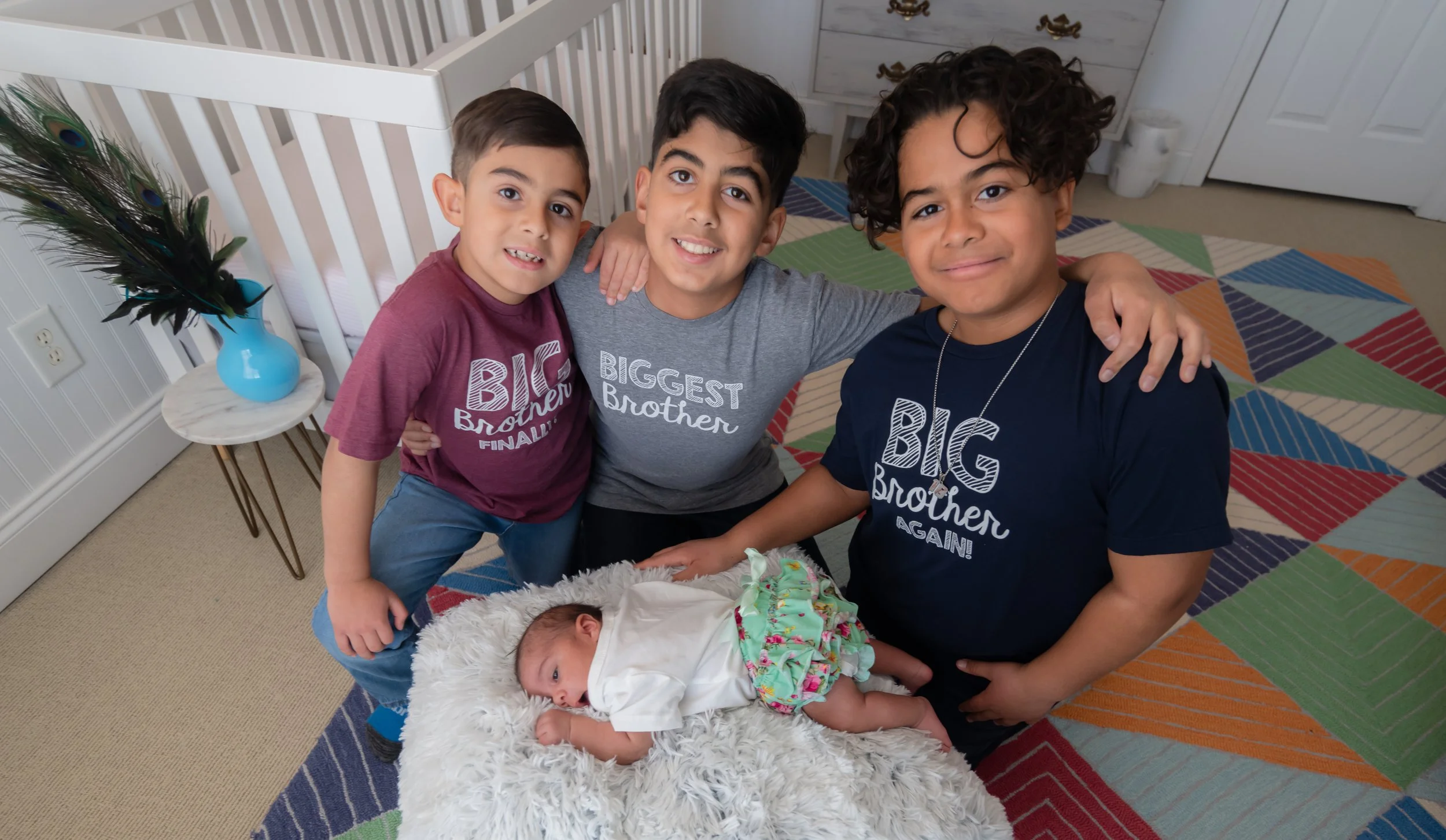Three young brothers with different hair colors and styles, wearing matching 'Big Brother' T-shirts, surround a newborn baby lying on a fluffy white blanket in a nursery with colorful geometric patterned rug, a white crib, and a small table with a va