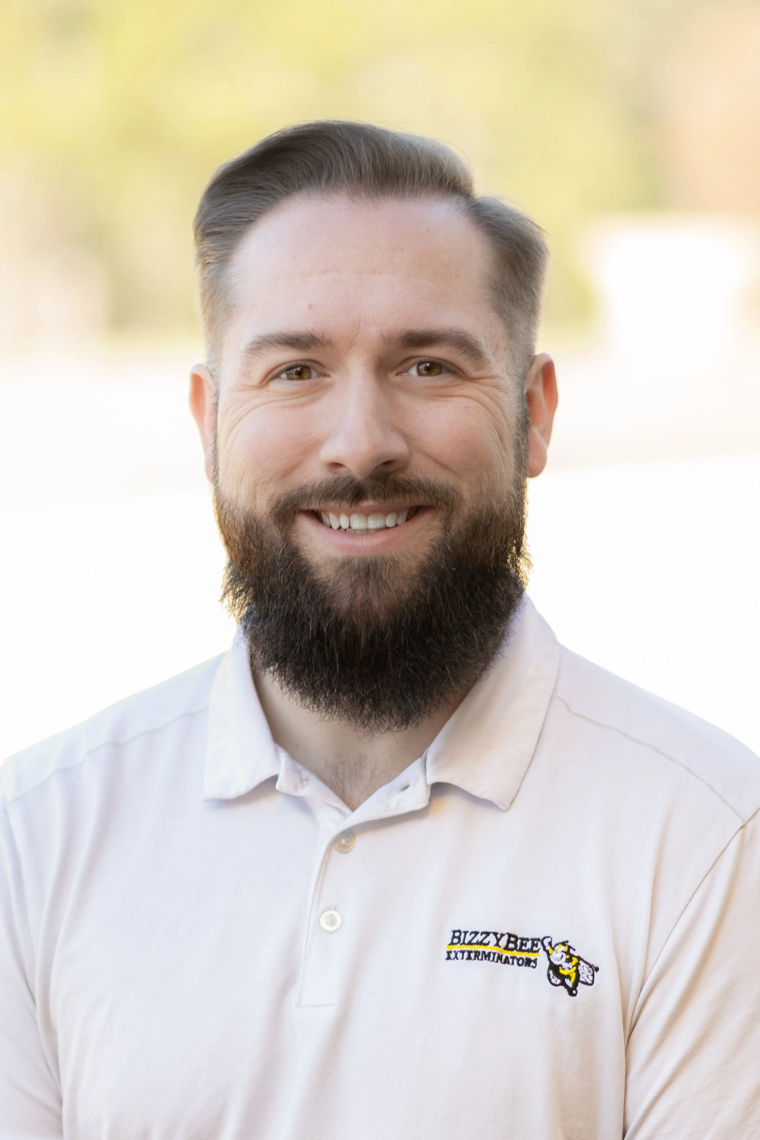 A smiling man with a beard and short styled hair, wearing a white polo shirt with a logo that says "BIZZY BEE EXTERMINATORS" with a cartoon bee holding a fly swatter, standing outdoors with blurred background of trees and sky.