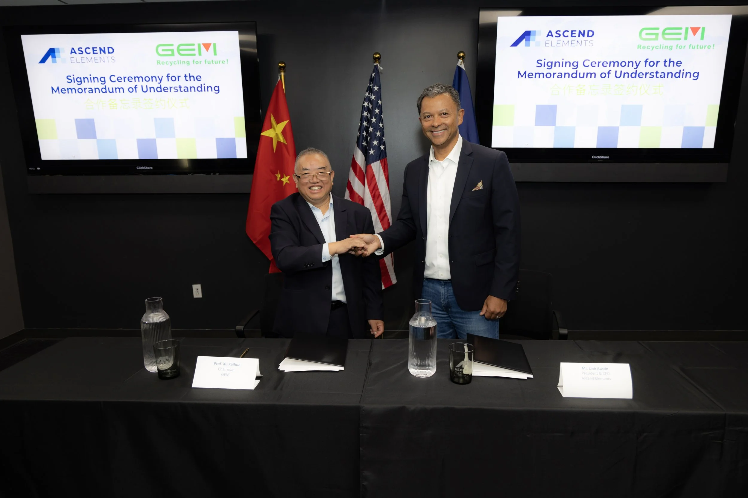 Two men in suits shaking hands in front of screens displaying logos and text about a signing ceremony for a Memorandum of Understanding. There are flags of China and the United States behind them, and a conference table with documents, water pitchers