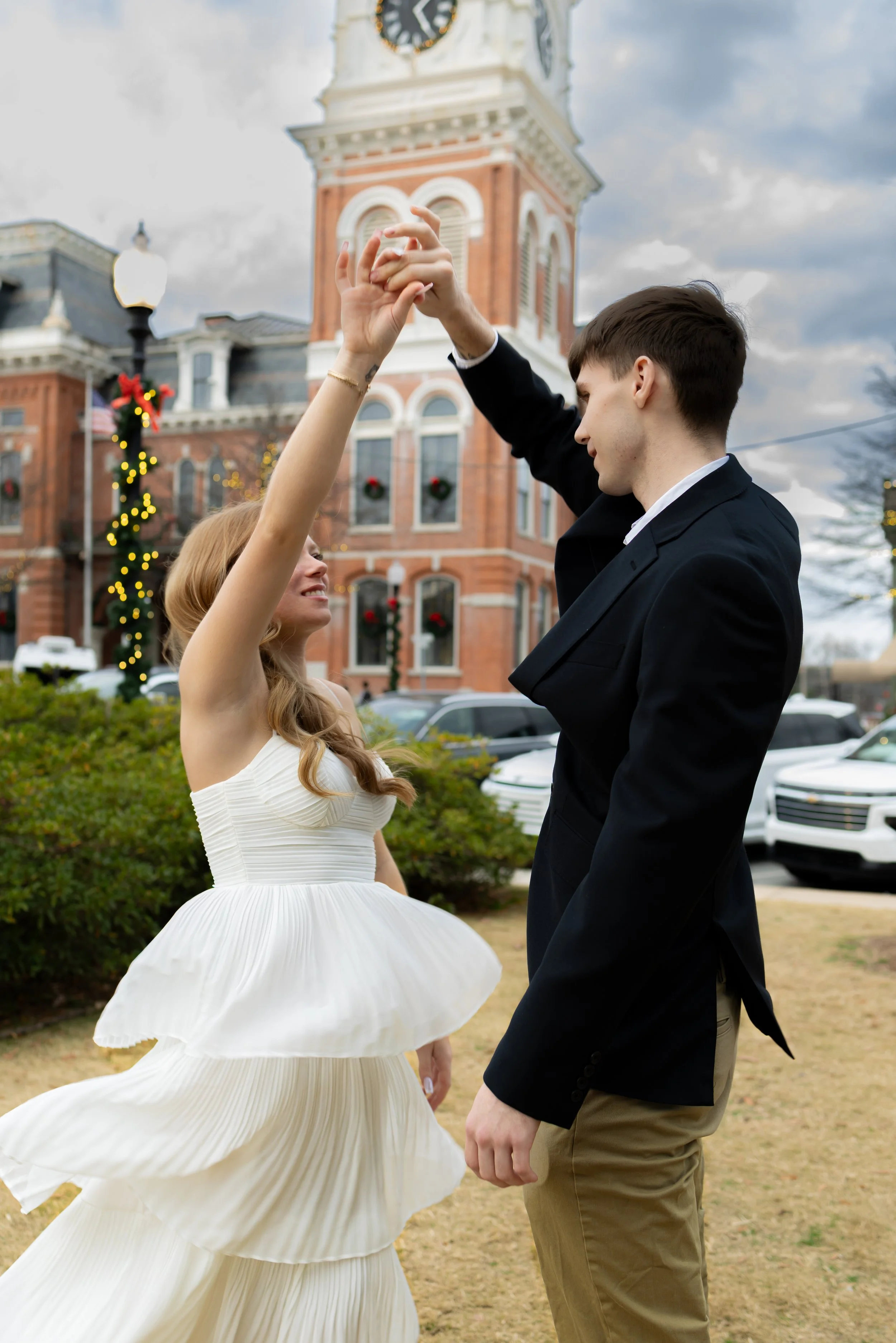 A couple dancing outdoors near a historic building decorated with Christmas wreaths and lights, with a clock tower in the background.