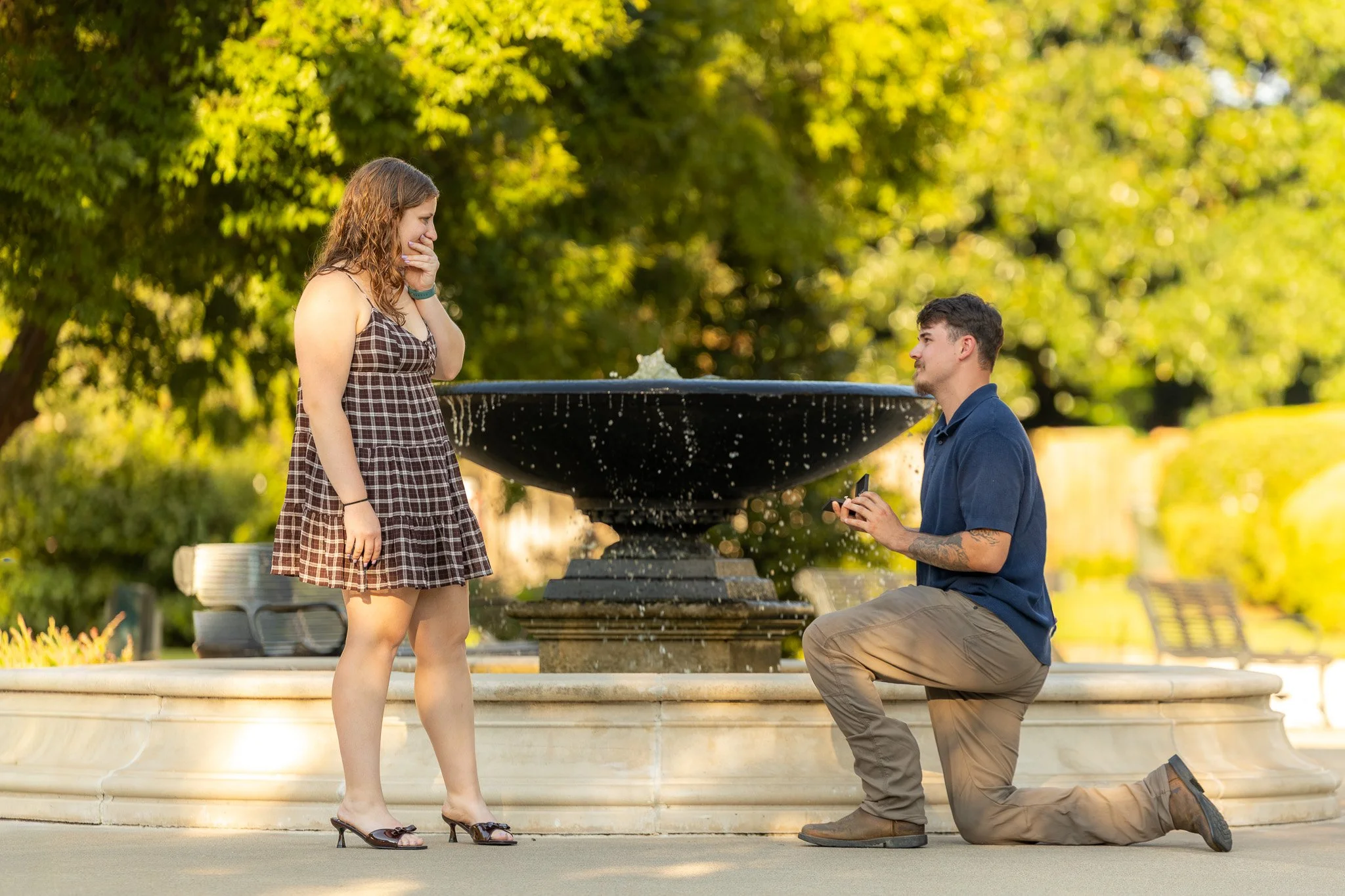 A man on one knee proposing marriage to a woman in a park with a fountain in the background.