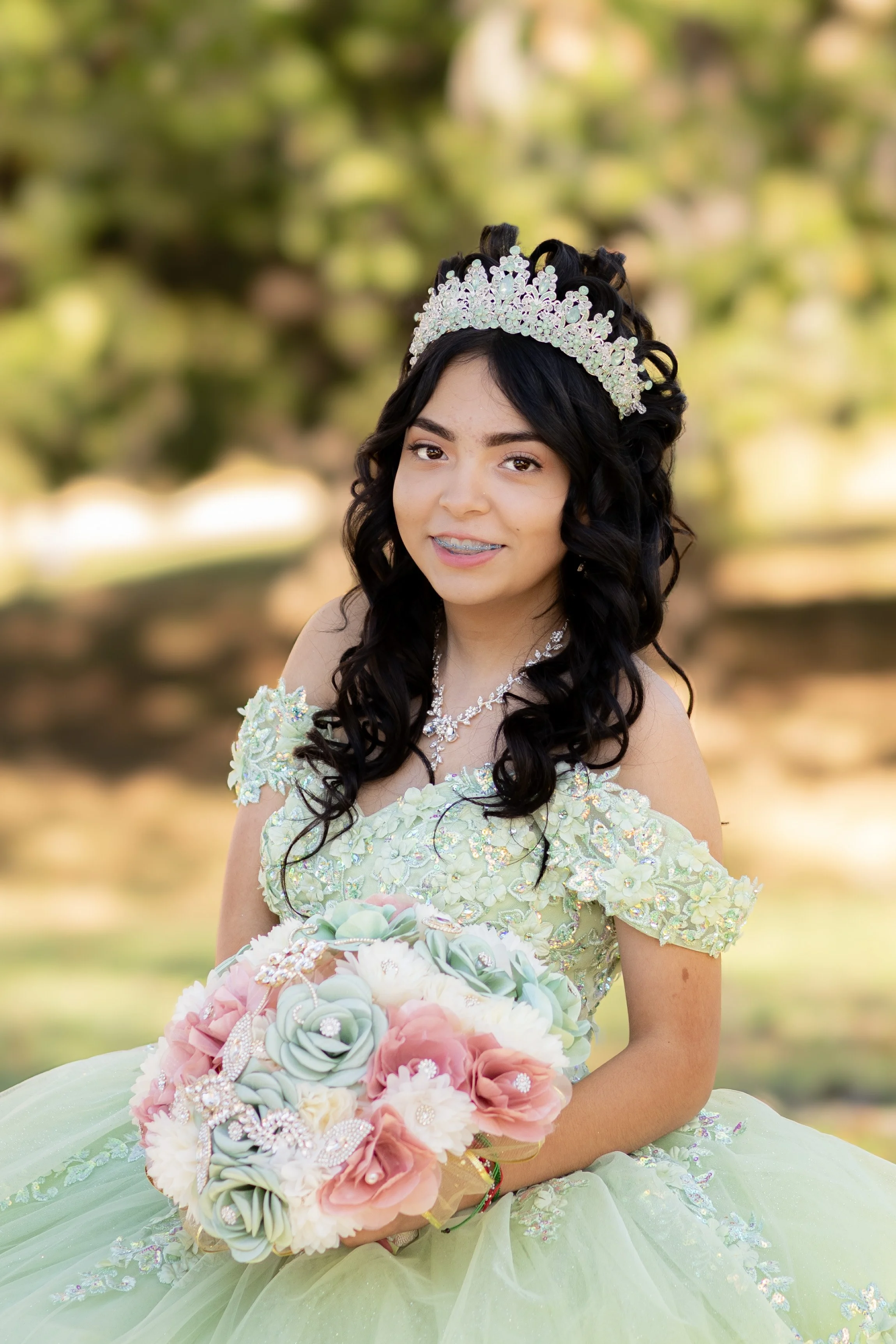 Young girl in a light green dress with floral embellishments, wearing a sparkly tiara and holding a bouquet of pastel-colored artificial flowers, outdoors with blurred trees in the background.