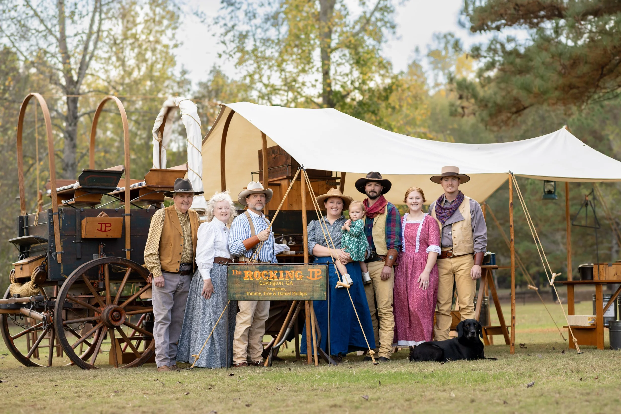 Group of people dressed in 1800s pioneer costumes standing in front of a covered wagon and tent outdoors, with trees in the background.