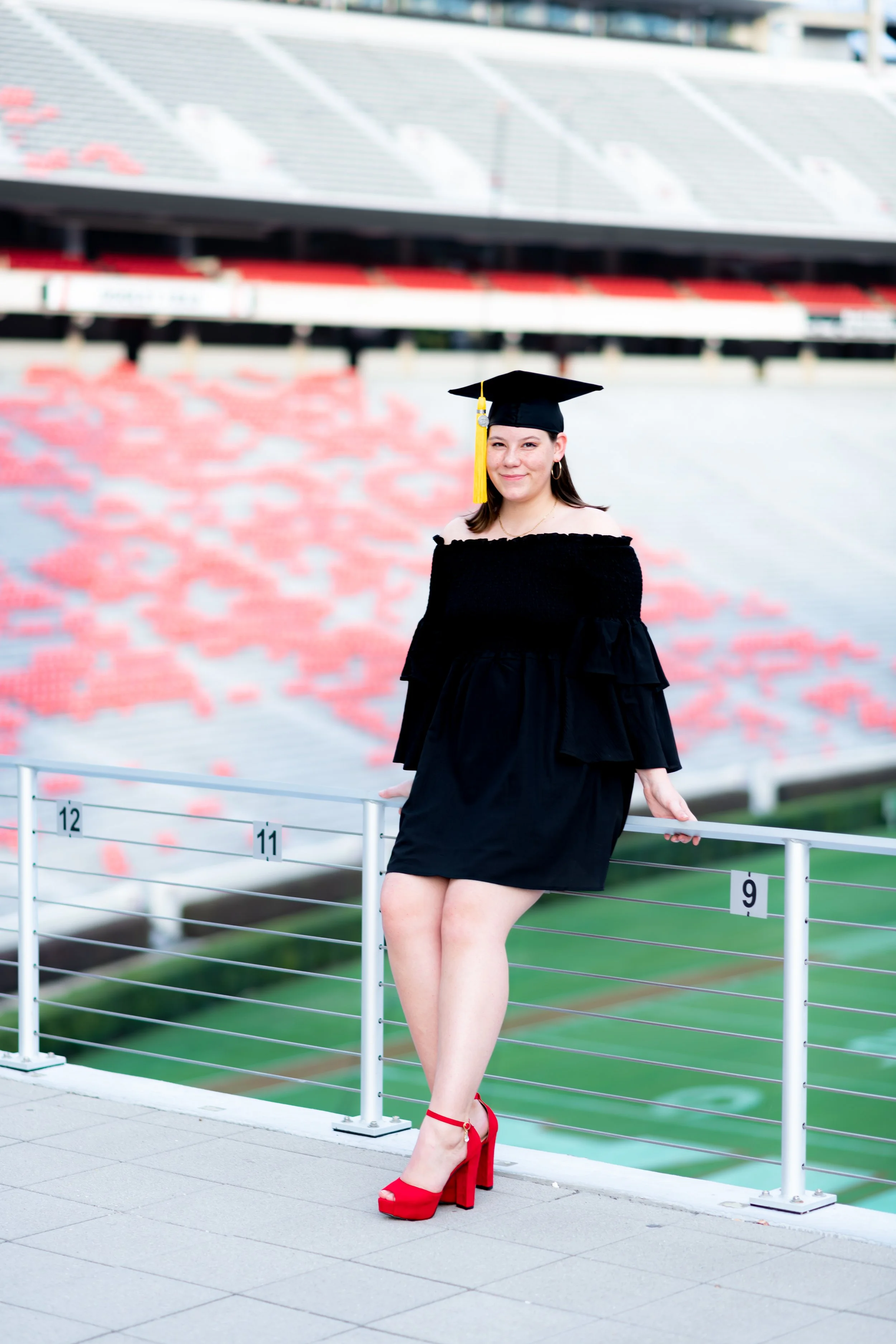 A young woman in a black graduation gown and cap with yellow tassel standing on a UGA stadium balcony, smiling at camera, wearing red high heels.