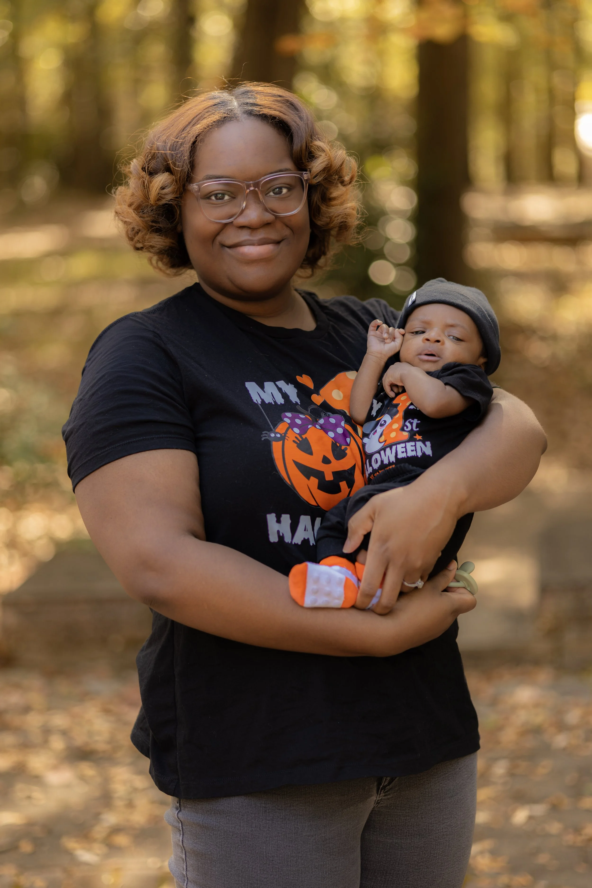 A woman holding a baby outdoors in a wooded area during autumn. The woman is wearing glasses and a black Halloween-themed shirt with a pumpkin design. The baby is dressed in a black outfit with Halloween decorations and a black beanie.