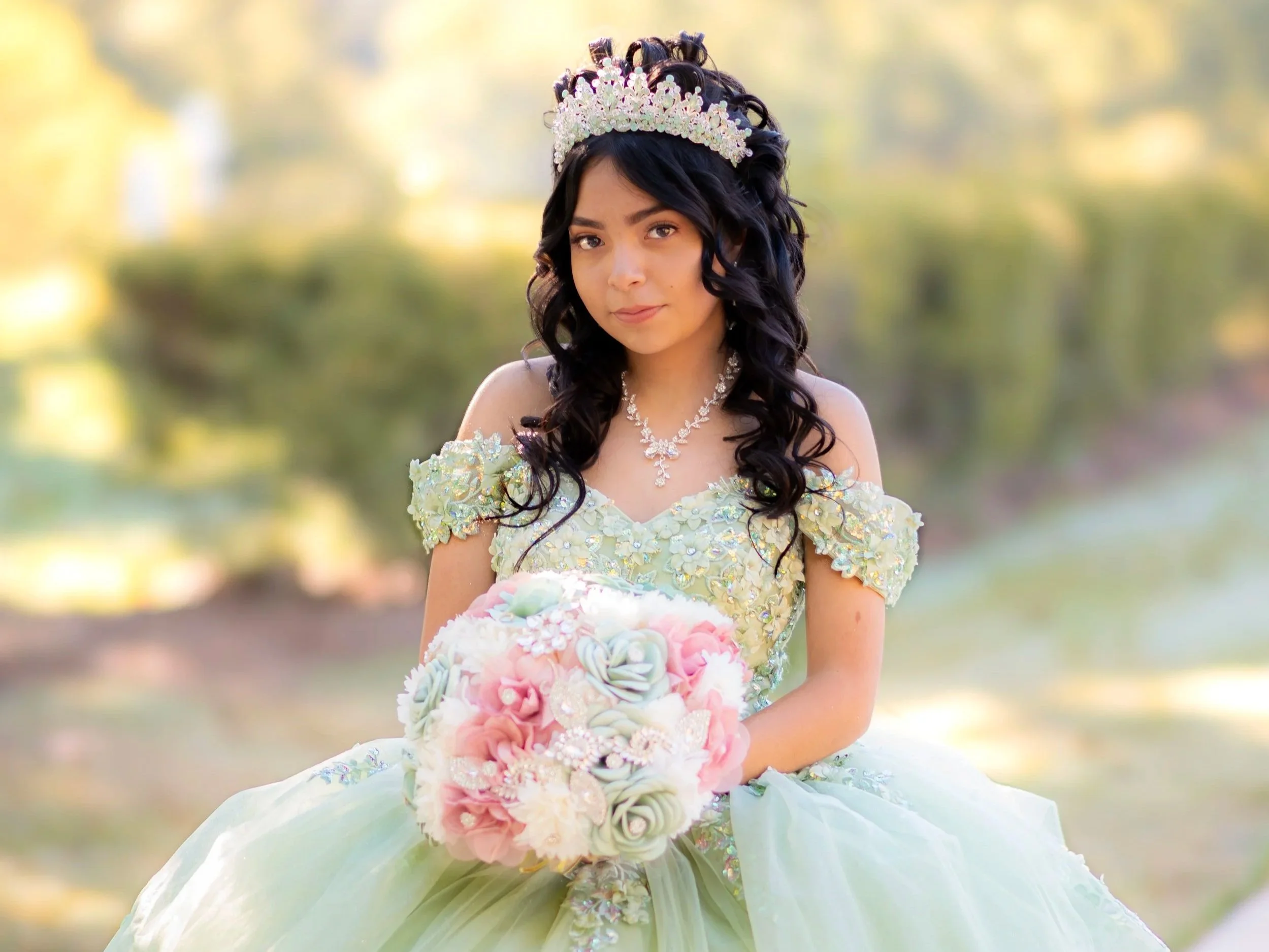 Young woman in a pastel green formal dress with floral accents, wearing a tiara and necklace, holding a bouquet of pink and pastel flowers, outdoors with blurred trees in the background.