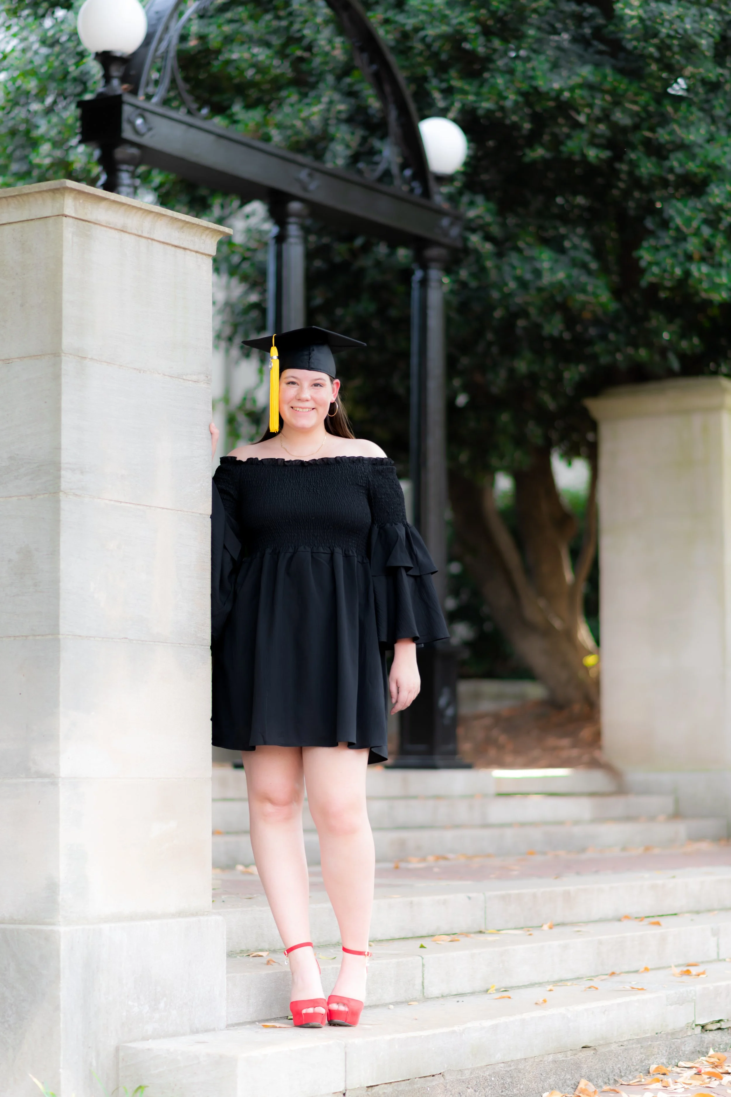 Young woman in a black off-the-shoulder dress and graduation cap standing outdoors on steps, smiling at camera.
