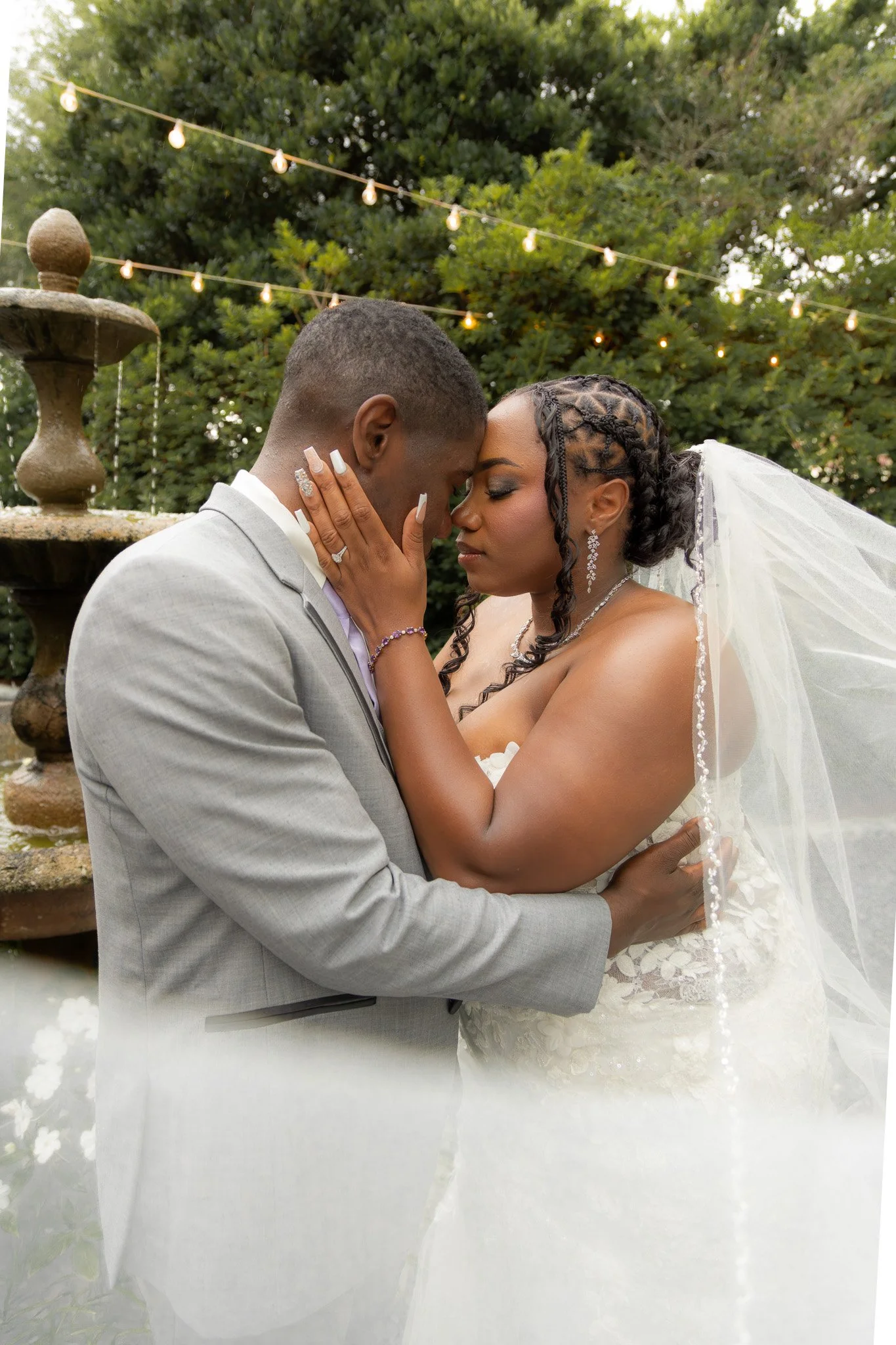 A newlywed couple dressed in wedding attire sharing an intimate moment behind a fountain and string lights in a garden setting.