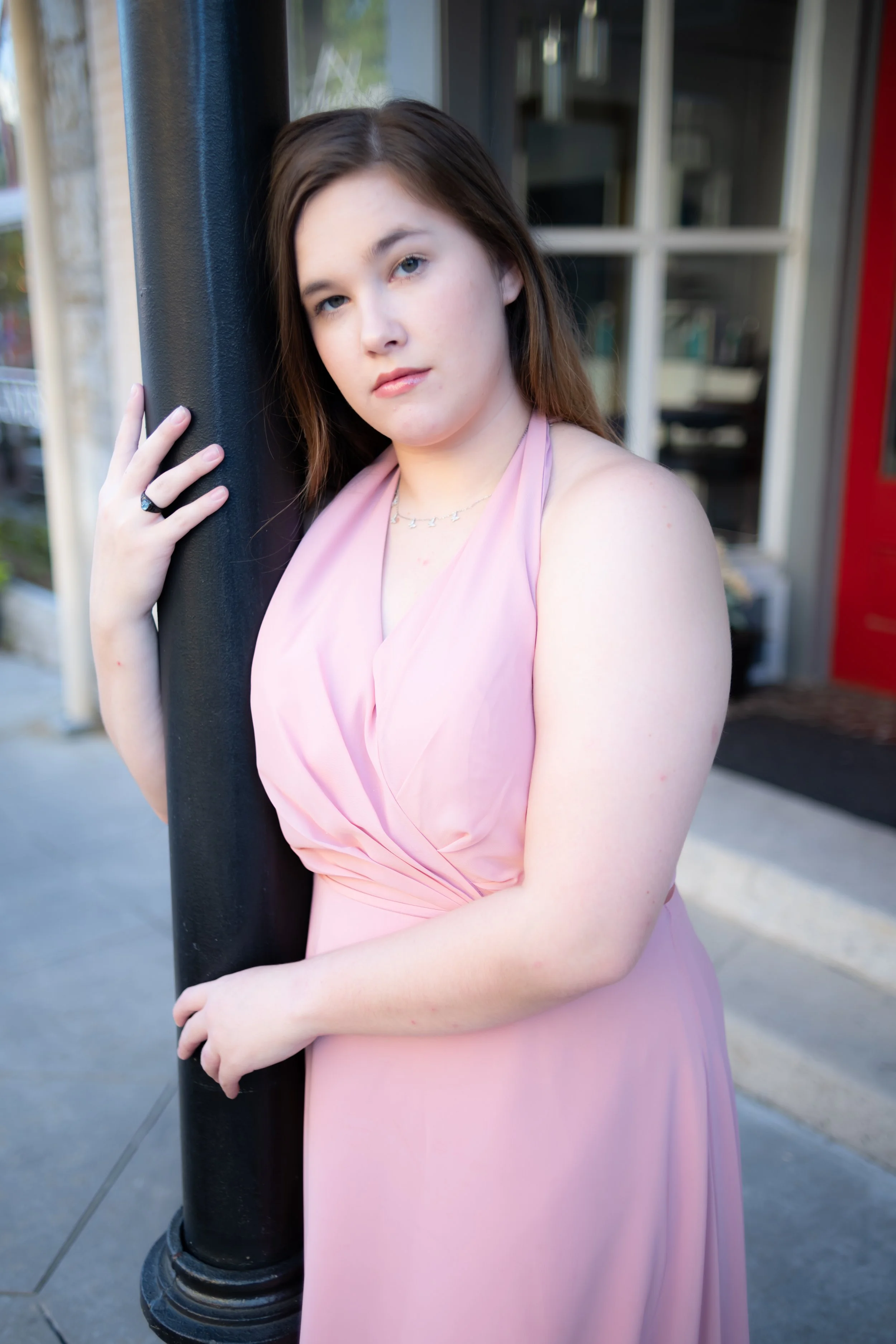 A young woman in a pink dress leaning against a black pole outdoors, with a building and window in the background.