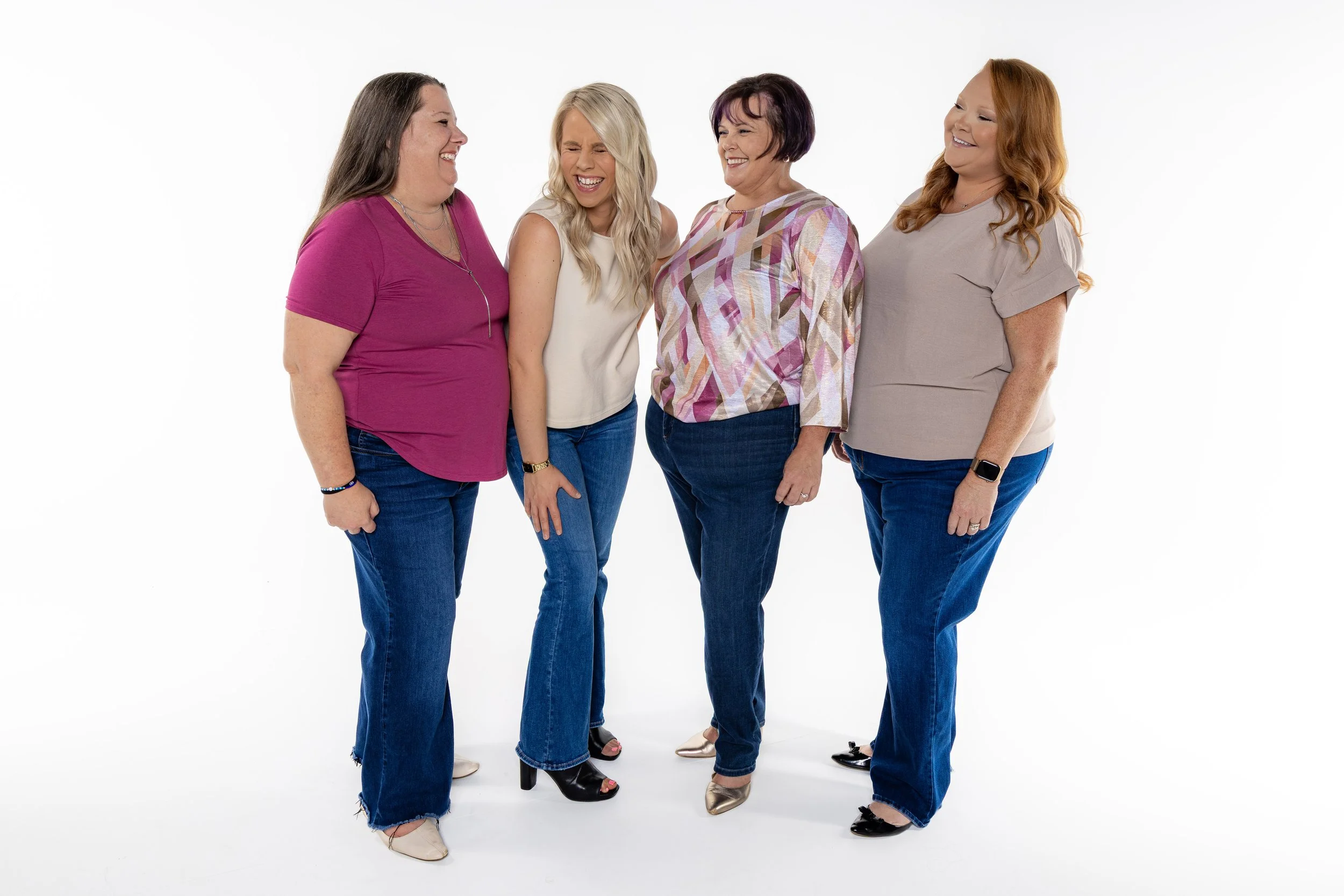 Four women standing together, laughing and smiling, in casual clothing against a white background.