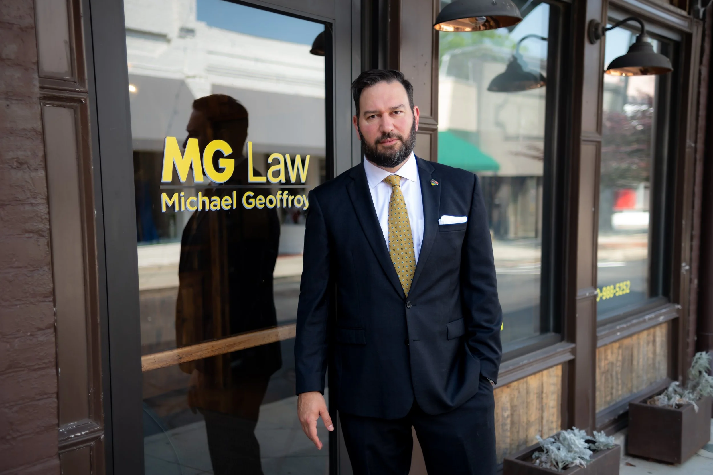 Man in navy suit with yellow tie standing in front of law firm office door with sign reading "MG Law Michael Geoffroy"