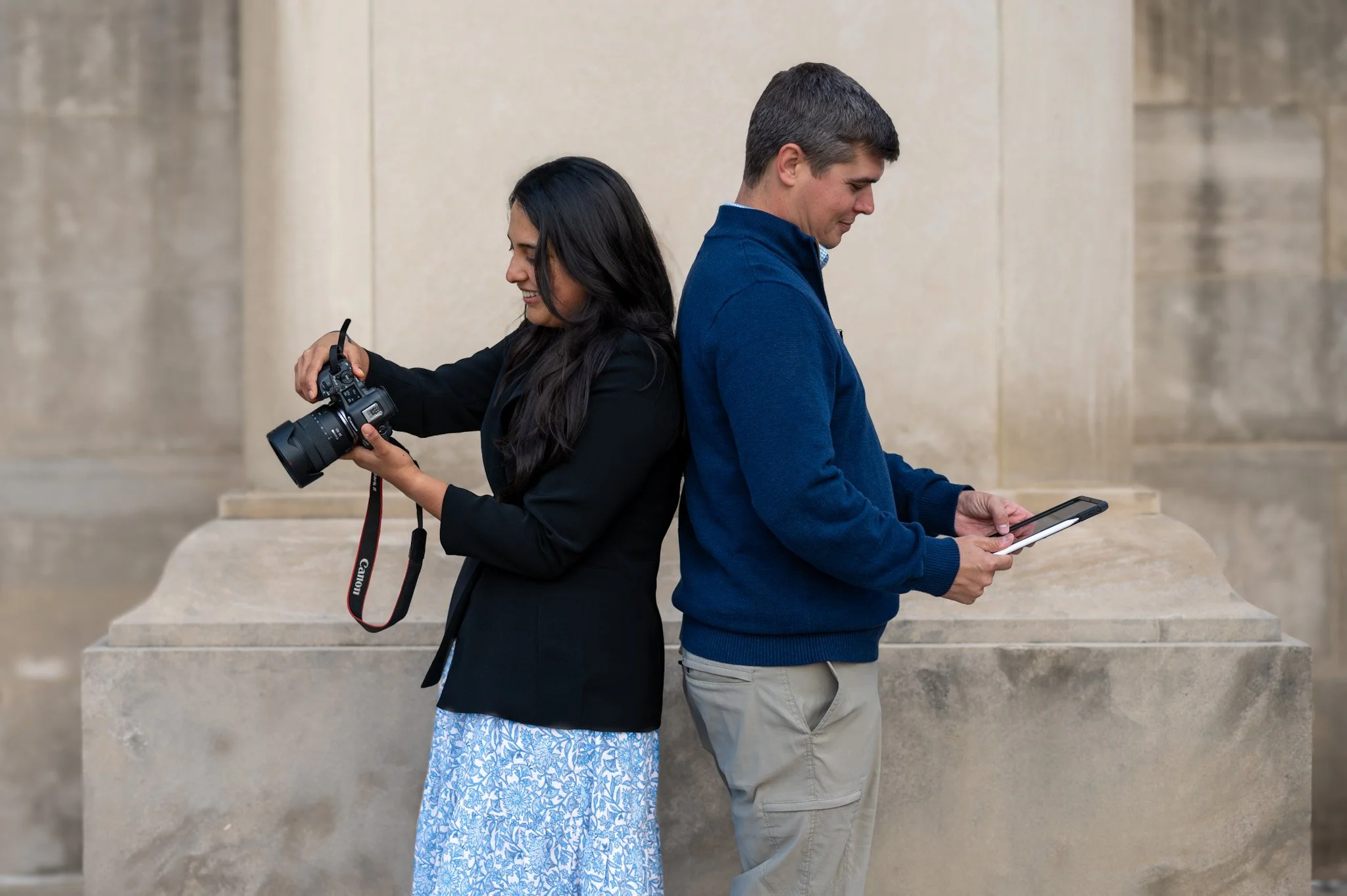 A woman holding a camera and a man looking at a tablet, standing back to back outside a stone building.
