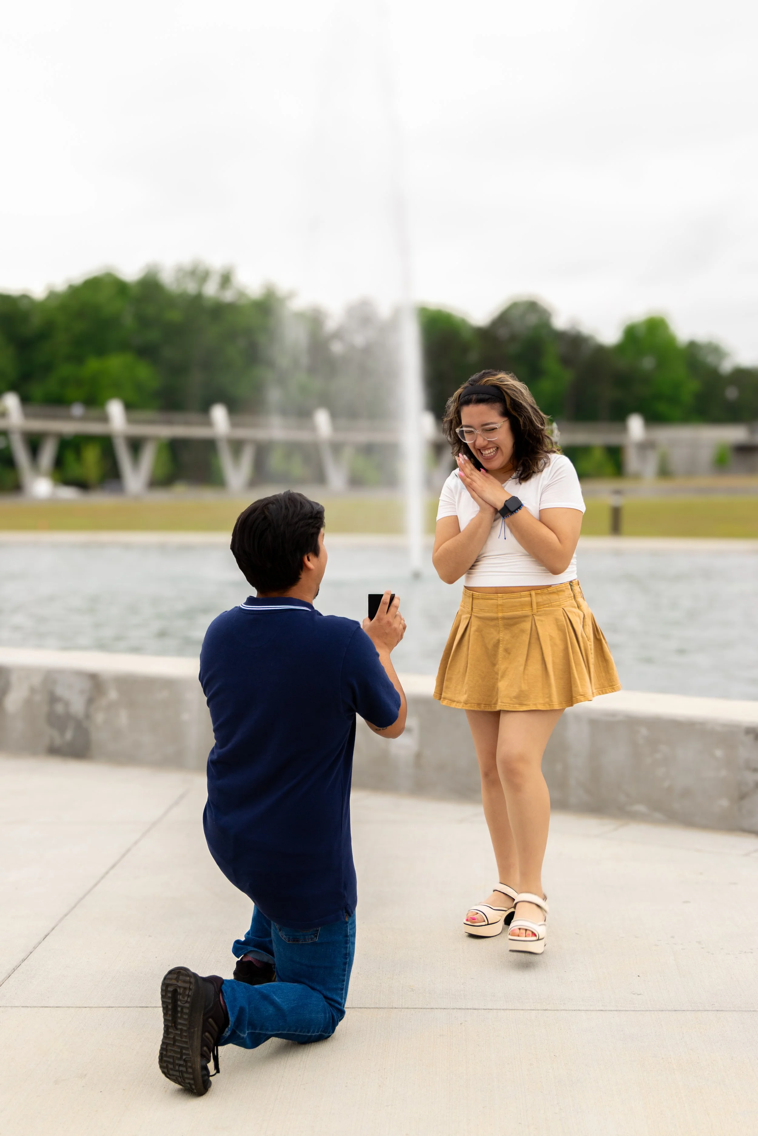 A man kneeling on one knee and proposing to a woman at a park with a fountain in the background. The woman is smiling and covering her mouth with her hands.