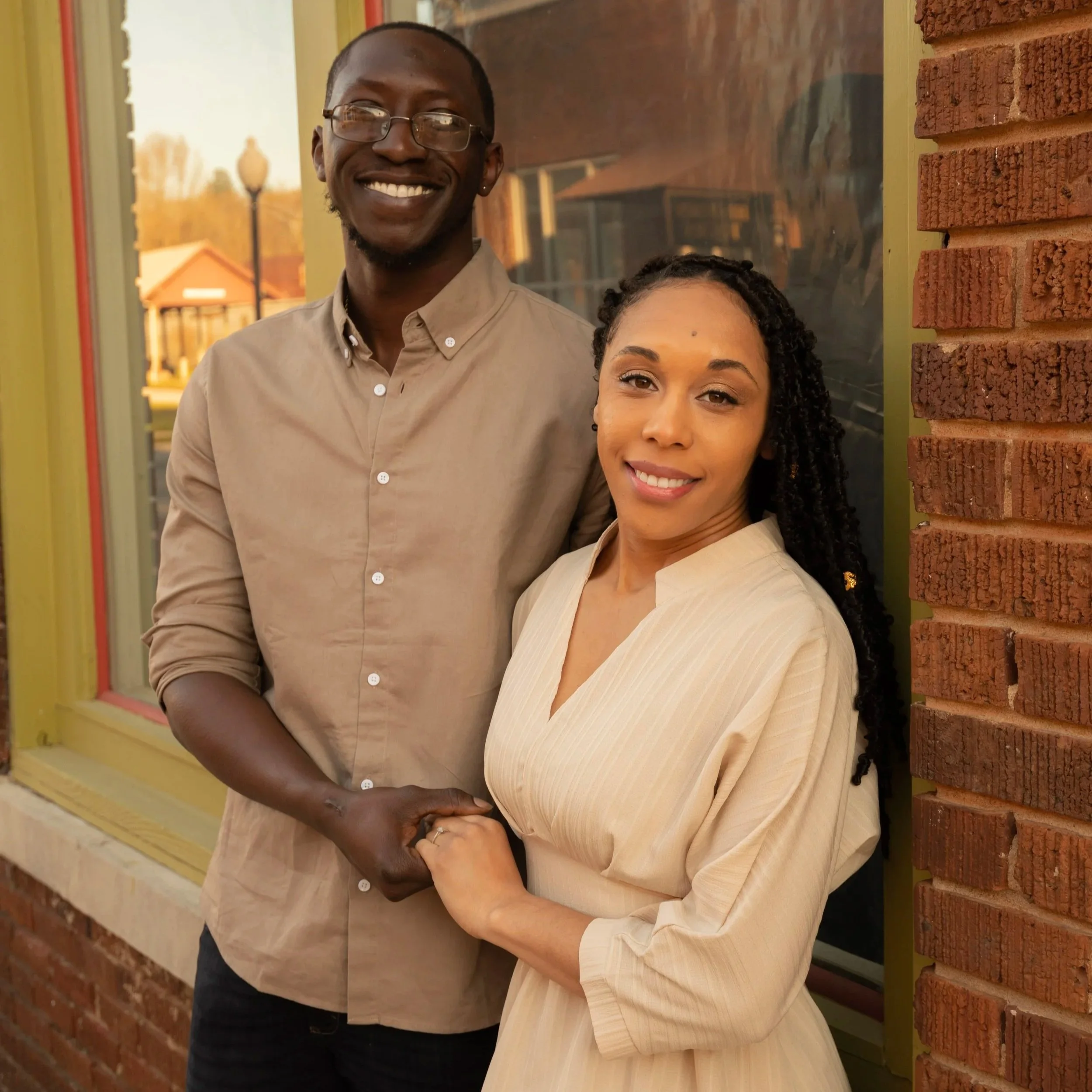 A smiling couple holding hands outside a building with brick walls and a window, with trees and buildings in the background.