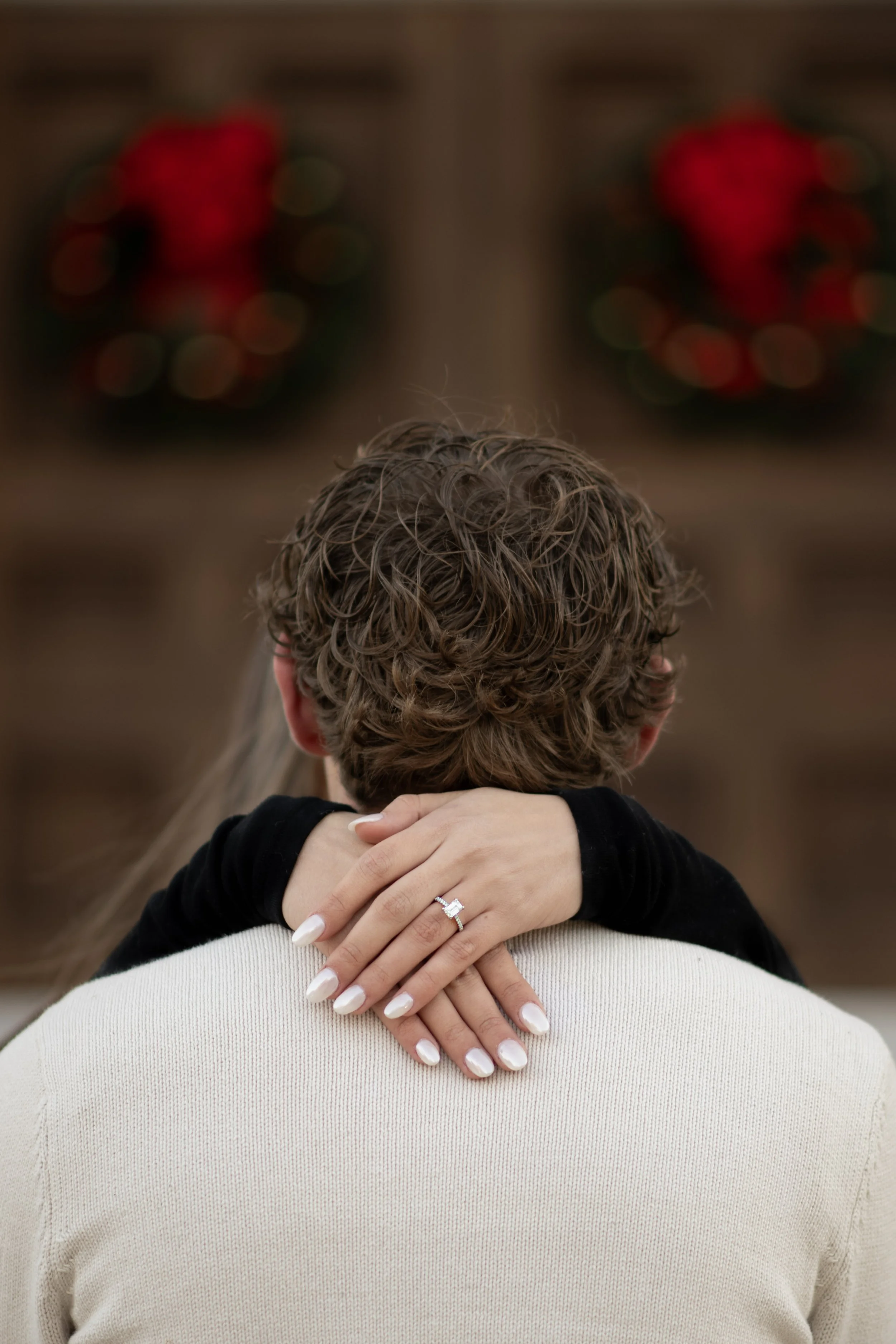 A woman with manicured nails and an engagement ring embraces a man with curly hair, with Christmas wreaths hanging in a wooden wall in the background.