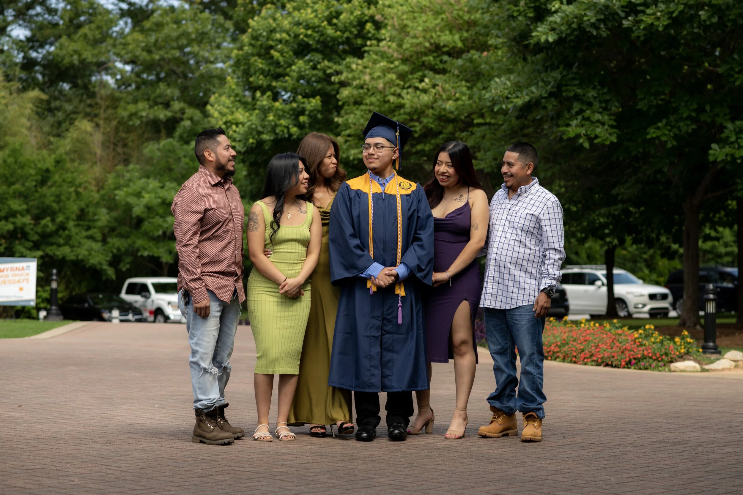 A young man in a blue graduation cap and gown with gold accents, standing on a brick walkway, surrounded by five people, with trees and parked cars in the background.