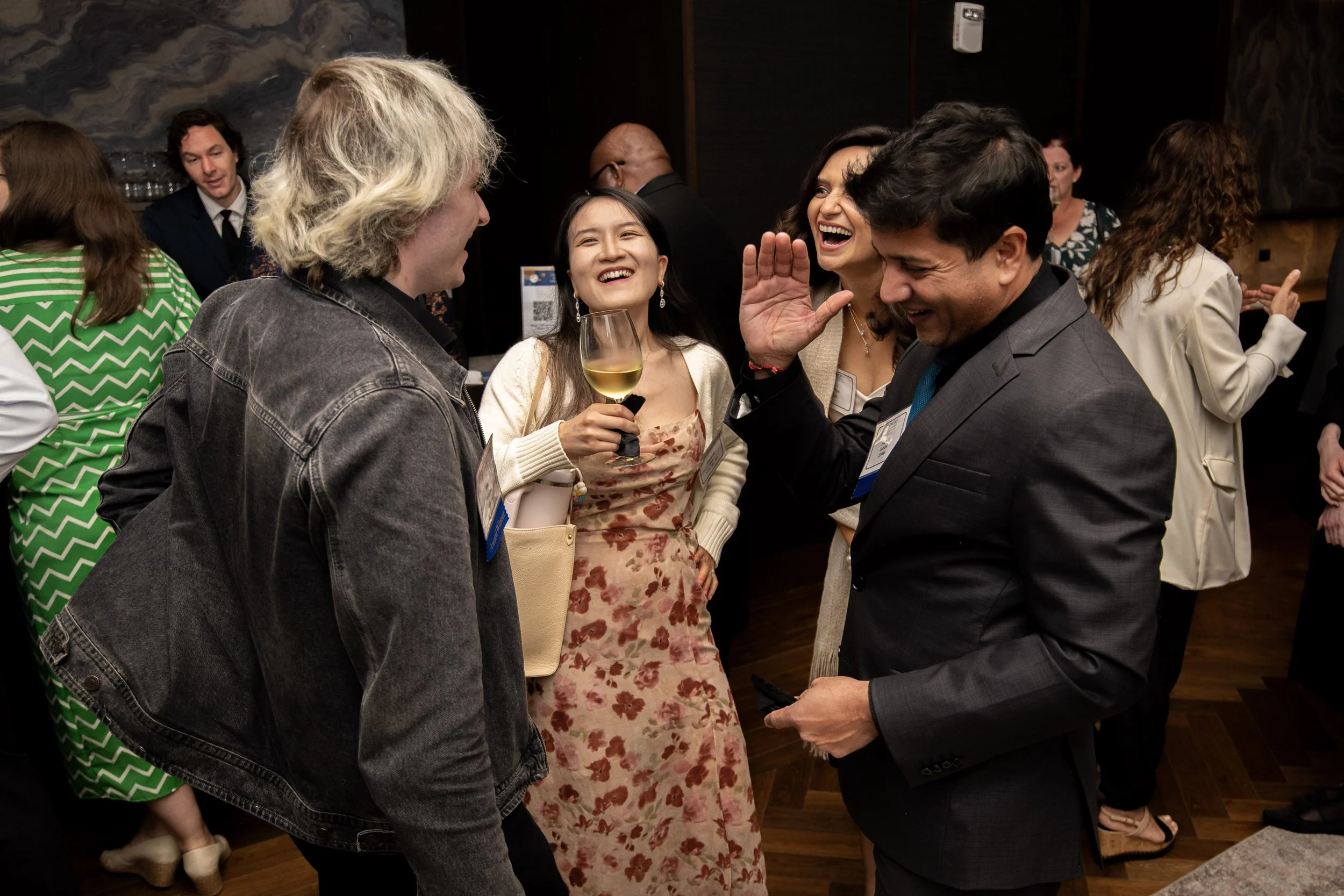 People socializing at a gathering, smiling and laughing, with some holding drinks, in an indoor setting.