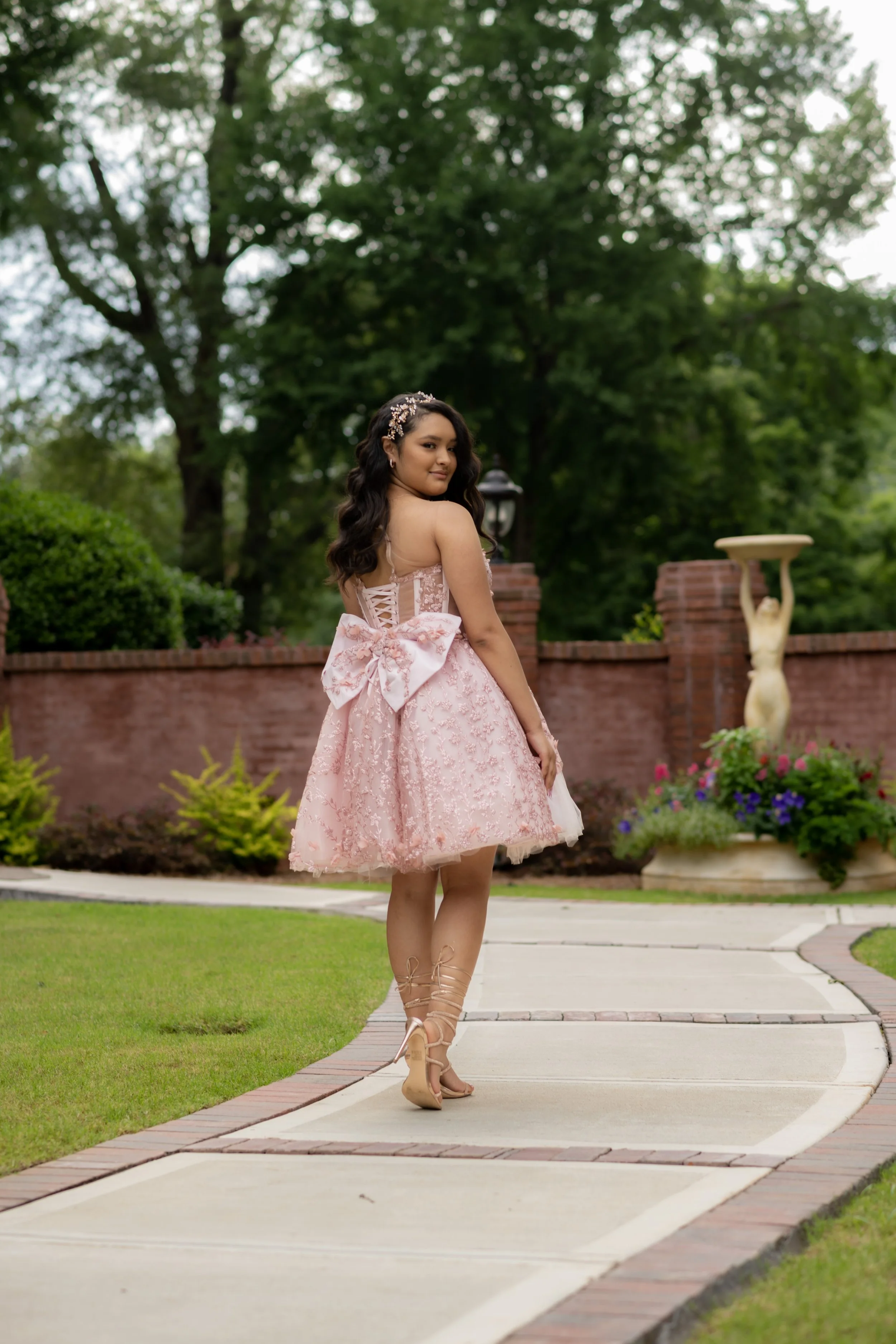 Young woman in a pink lace dress with a large bow on the back, standing on a curved sidewalk in a garden, looking over her shoulder, with trees and a brick wall in the background.